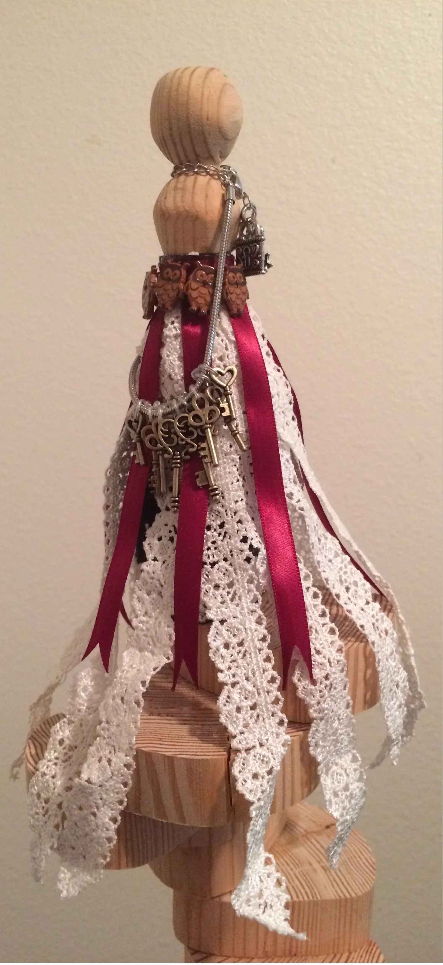 A decorative doll with a wooden head and neck, dressed in a vintage lace and ribbon dress, adorned with chains, keys, and owl charms.
