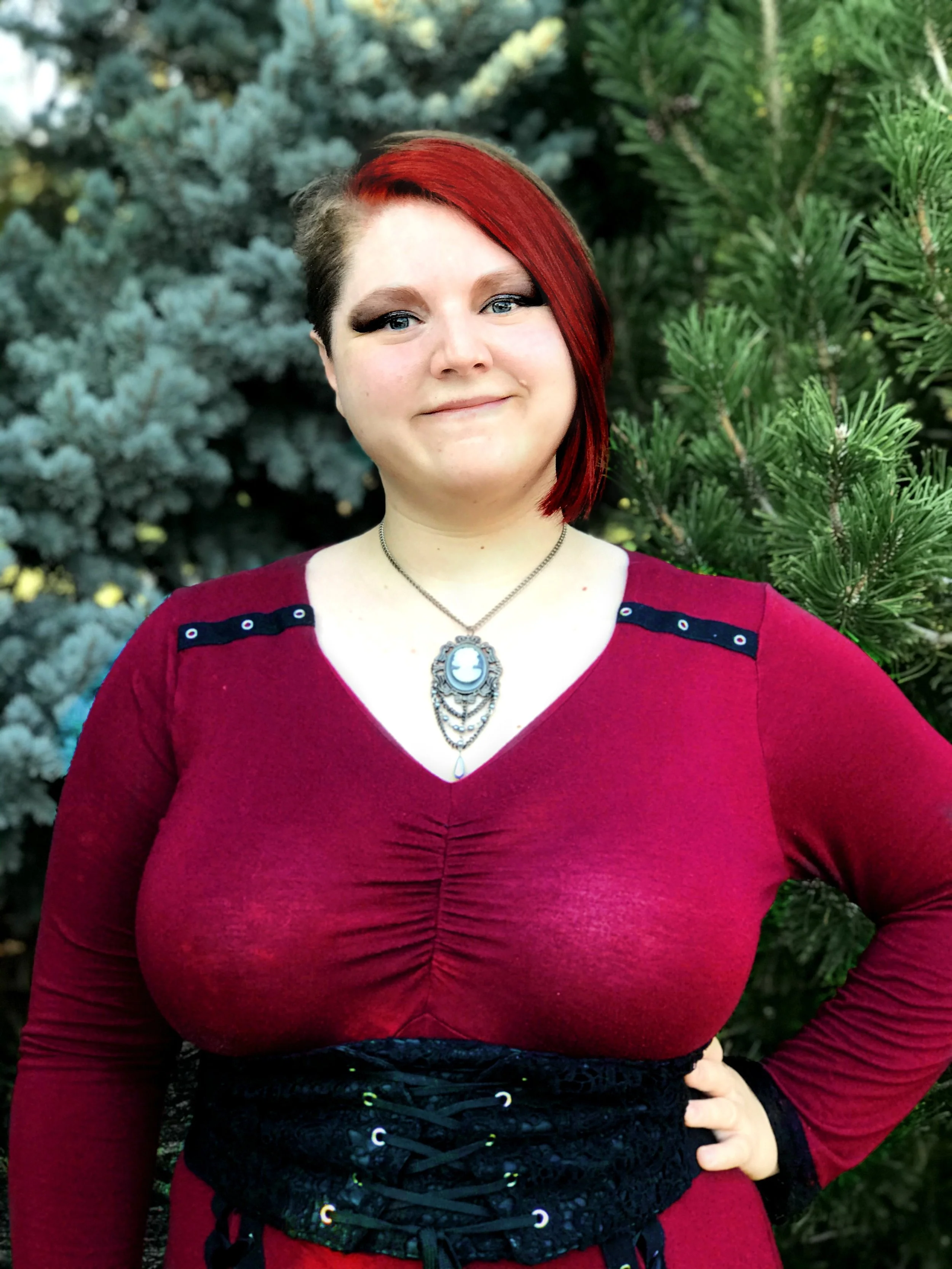 A woman with red hair styled in an asymmetrical cut, wearing a red dress with embellished belt, and a necklace with a cameo pendant, standing outdoors in front of evergreen trees.