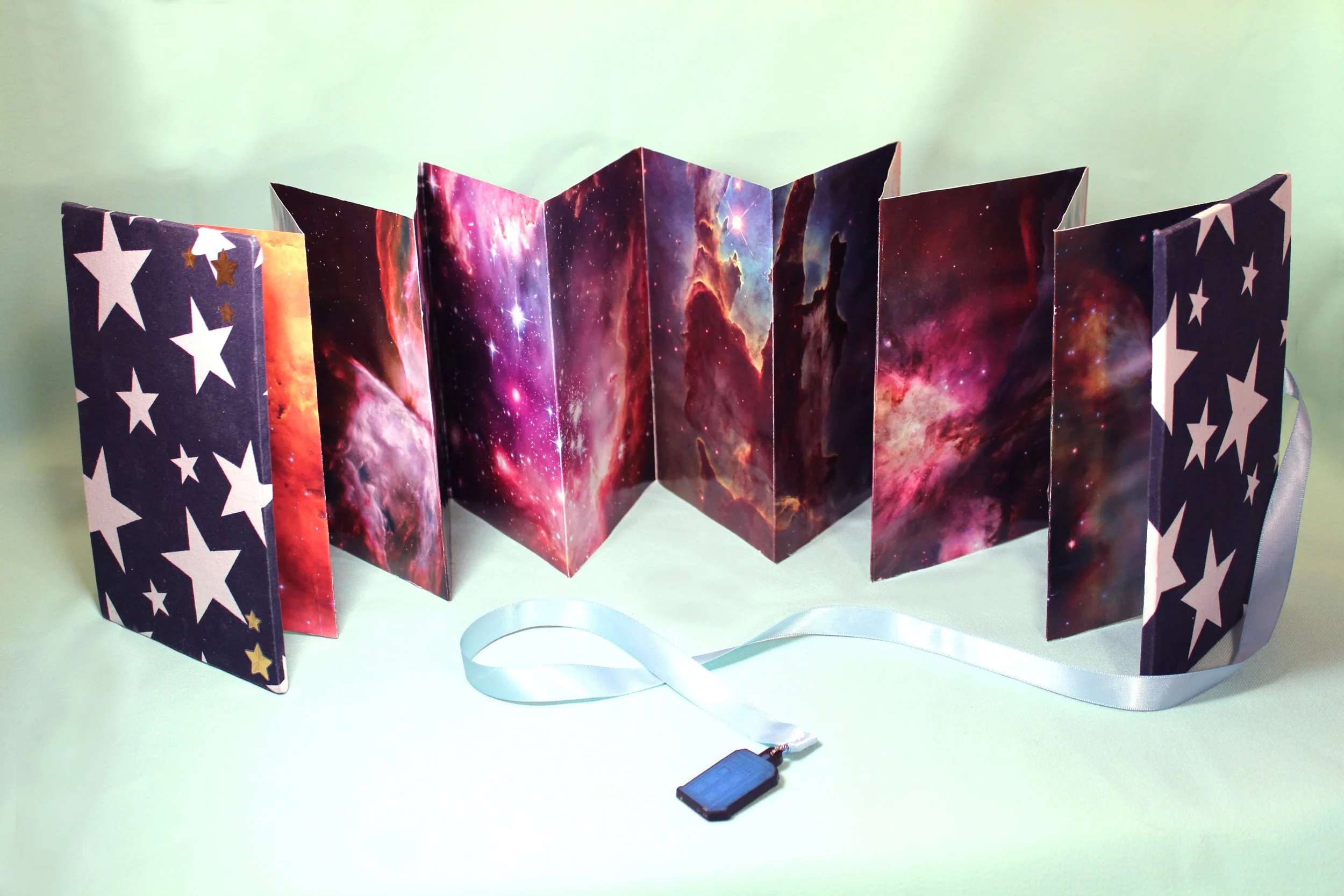 A folded space-themed paper display with galaxy imagery and star patterns on the sides, standing against a light green background, with a small electronic device or remote on the surface in front.