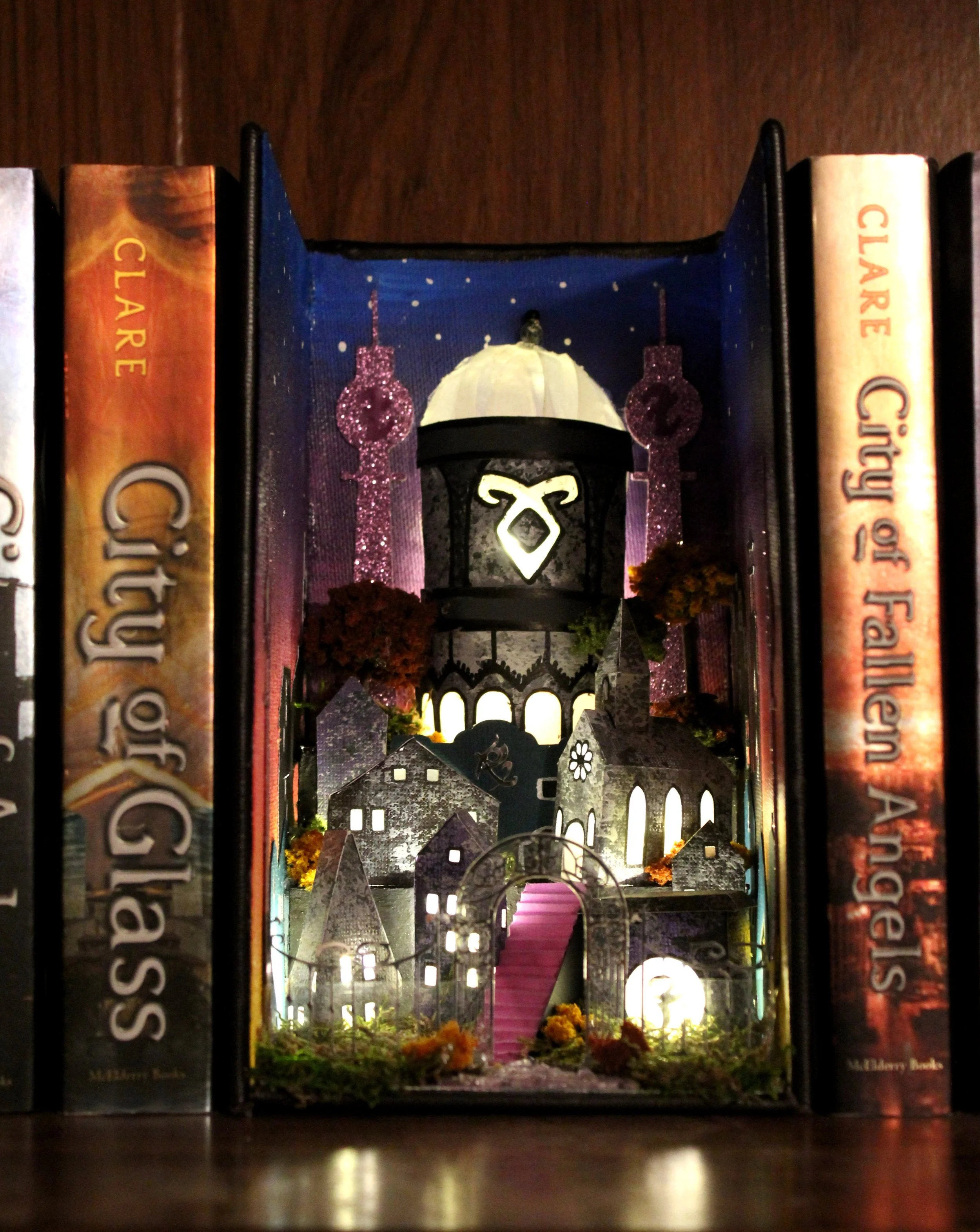 A miniature fairy tale village created with small buildings, a staircase, and a glowing lantern, viewed through a narrow opening between books on a shelf, with some evening flowers at the base.