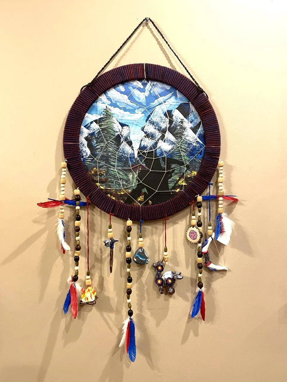 Circular dreamcatcher with a mountain landscape picture inside, decorated with beads, feathers, and small charms hanging from the bottom.