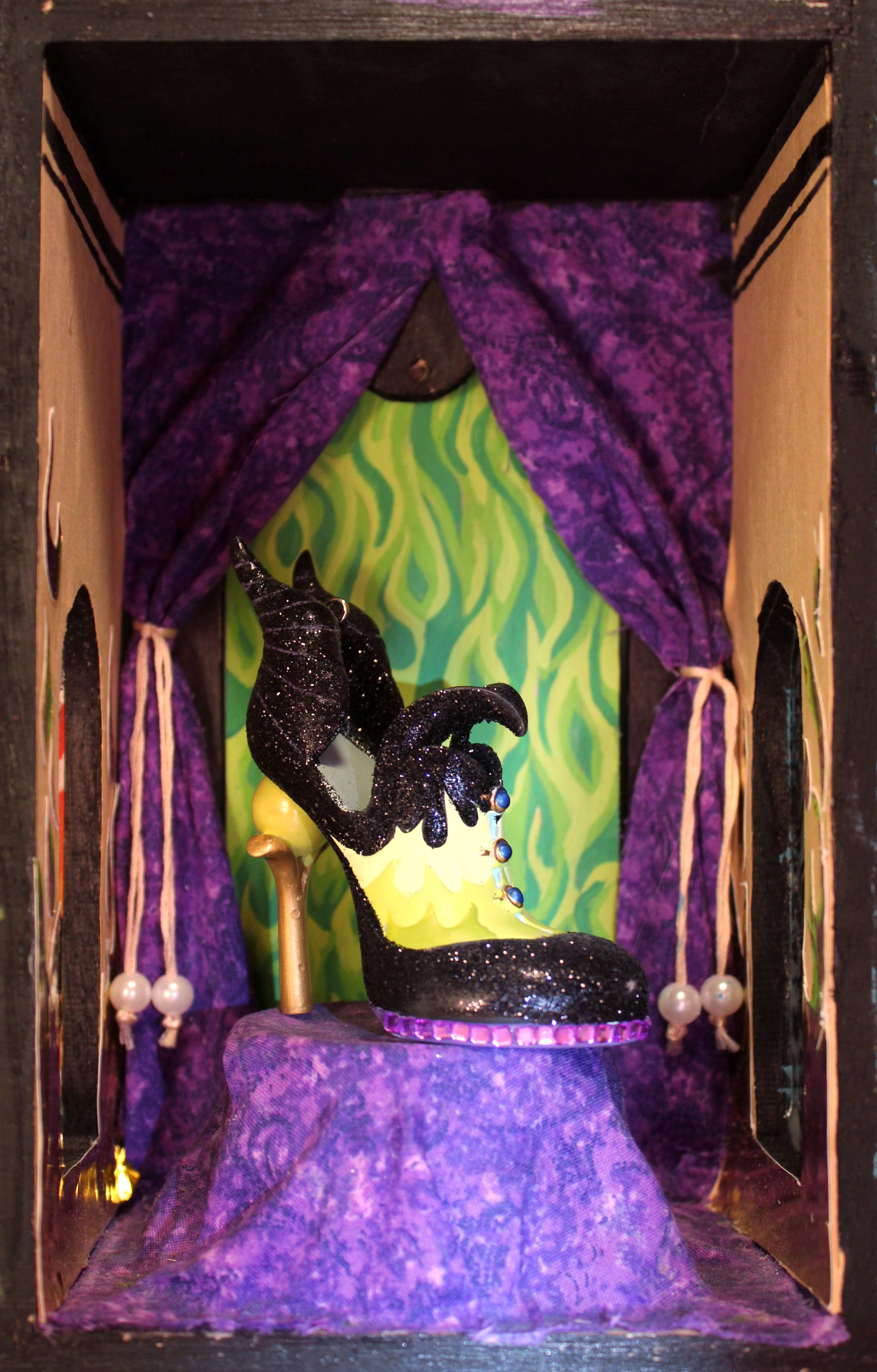 A miniature stage set featuring a black high-heeled shoe with purple glitter, a yellow heel, and blue gemstone accents, placed on a purple fabric with a green and yellow zigzag background, framed by purple velvet curtains with pearl tassels, inside a