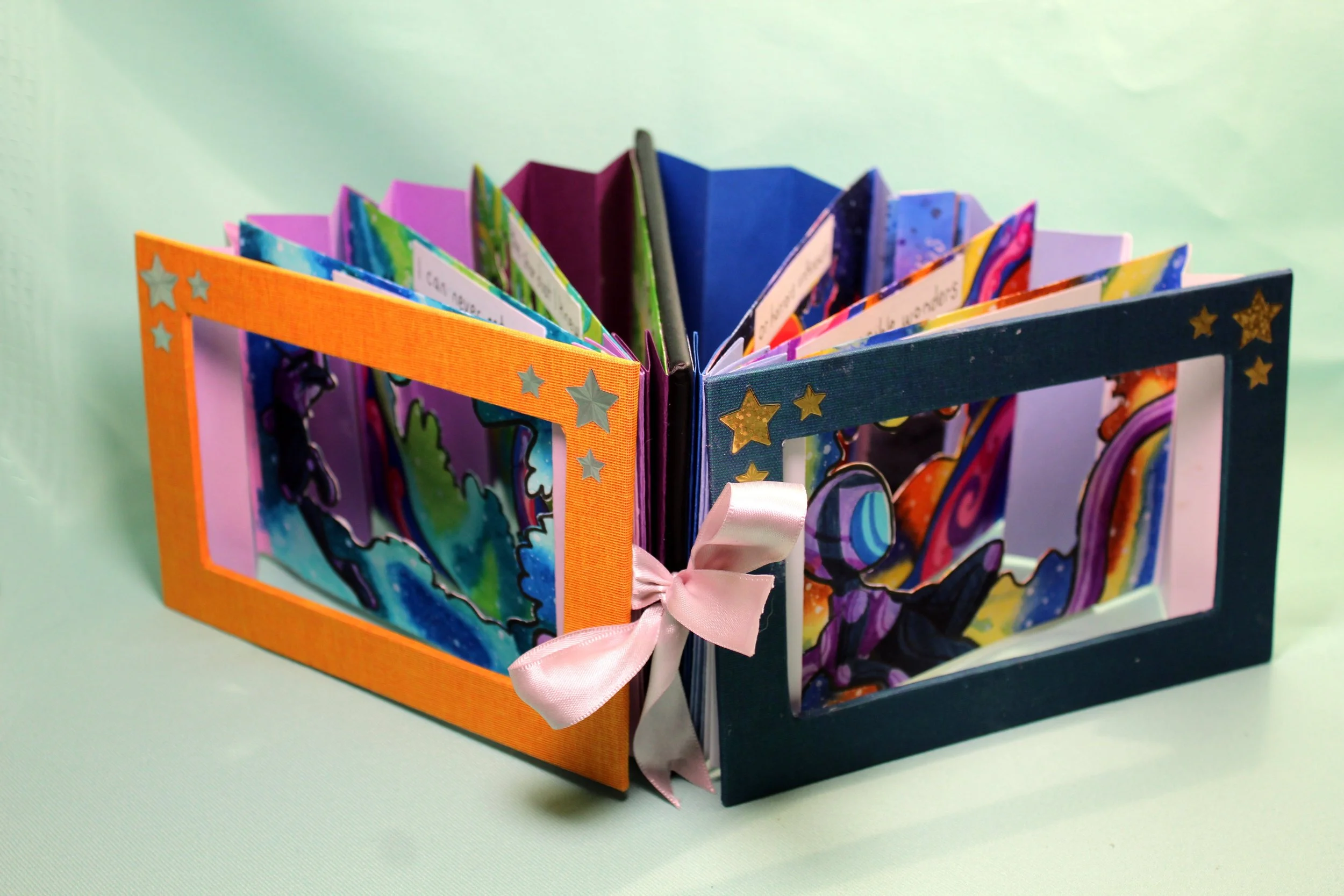 A colorful, handmade accordion-style flipbook with star decorations on the cover, tied with a pink ribbon, displayed on a pastel green background.