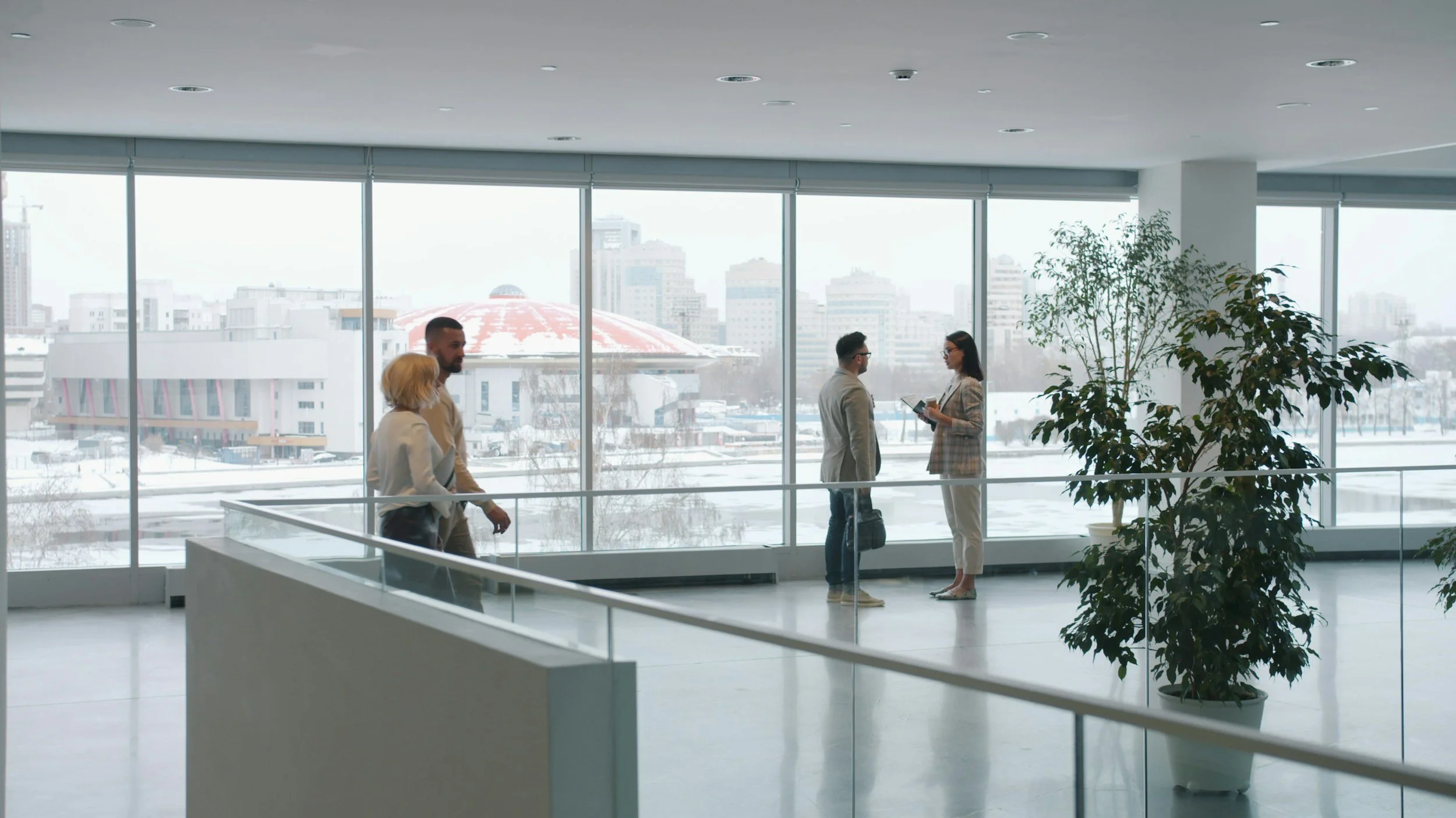 Four people in business attire standing and talking with each other near large windows in an office building. Two women and two men, with a city view in the background, including a distinctive building with a red and white dome. The office has a large potted plant and snowy weather outside.