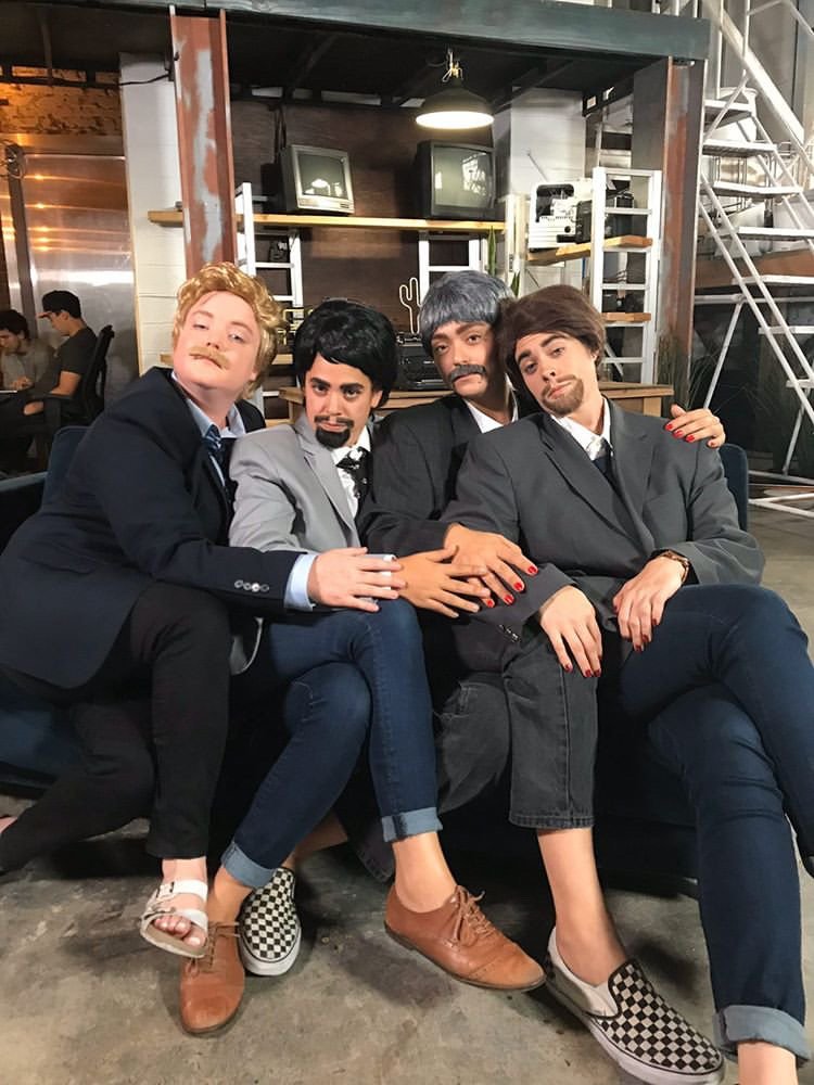 Four people sitting on a black couch dressed in suits, with mustache and beard makeup. They are posing for a photo in an industrial-style space, with shelves, TV screens, and a staircase in the background.