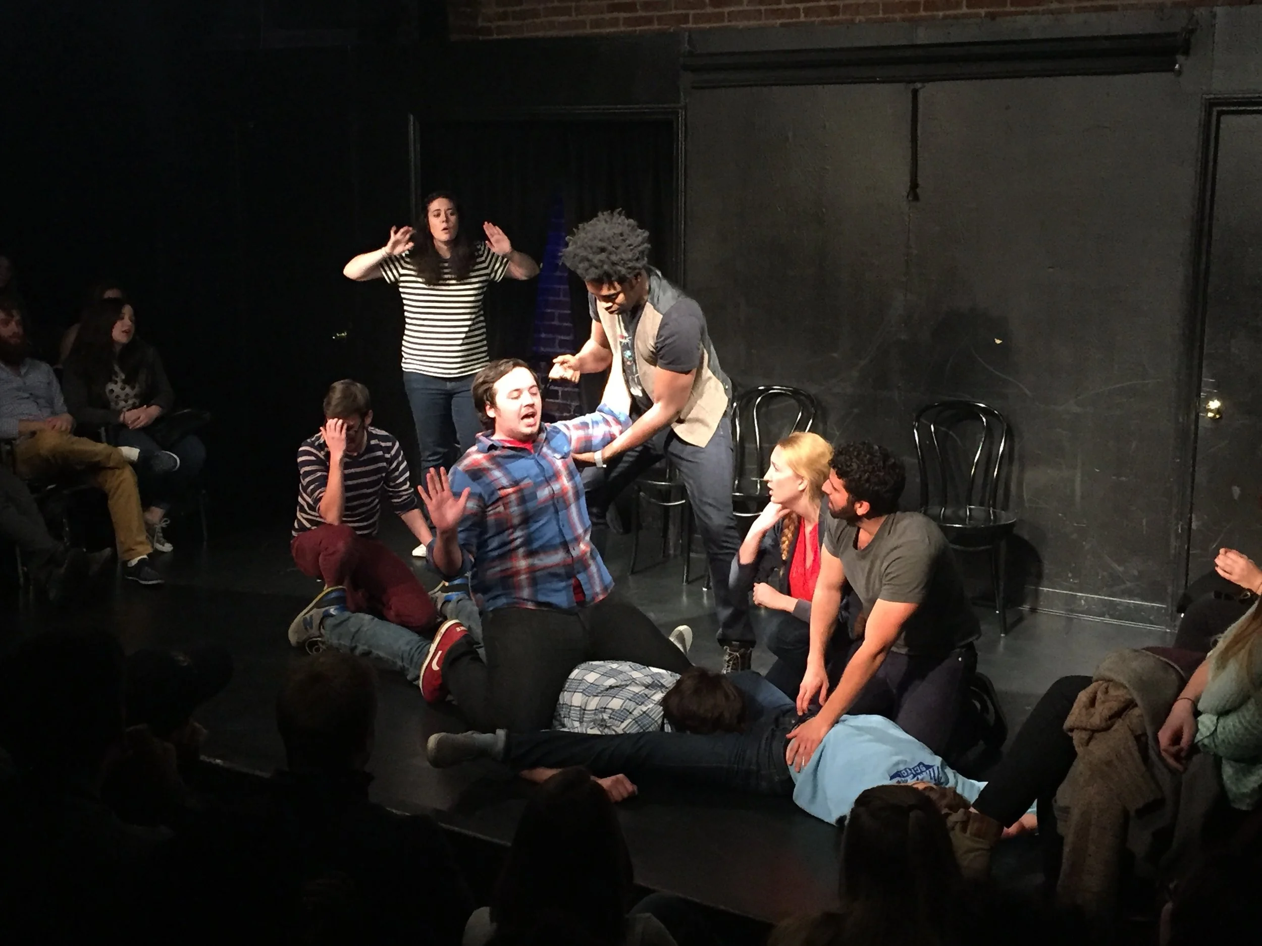 A group of people are performing a scene on stage with some seated and some standing. One man with curly hair and glasses is holding a woman lying on the floor, while another woman stands with her hands raised, and several audience members are watching.