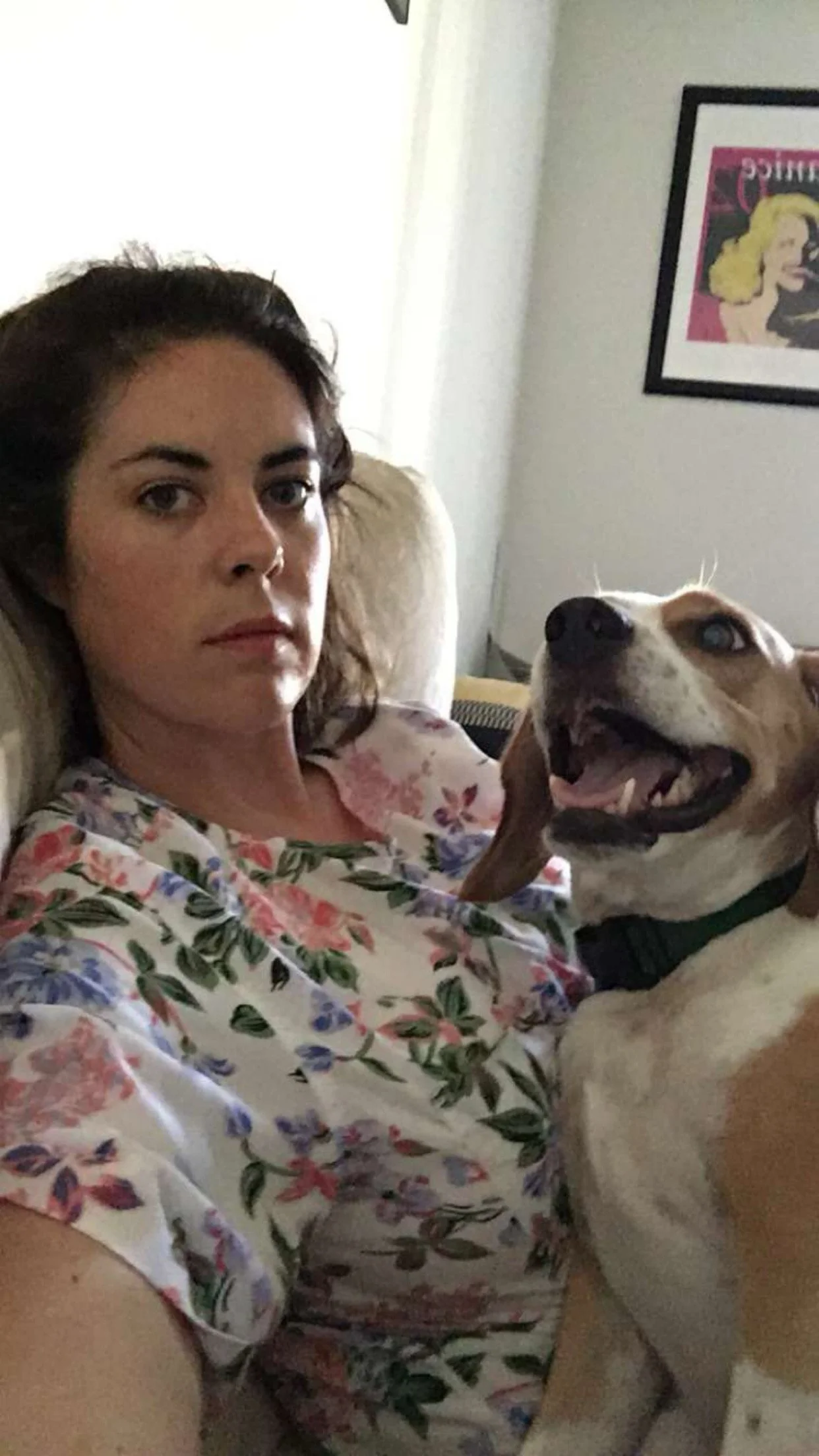 A woman with dark hair and a floral shirt is sitting on a couch with a smiling dog in her lap.
