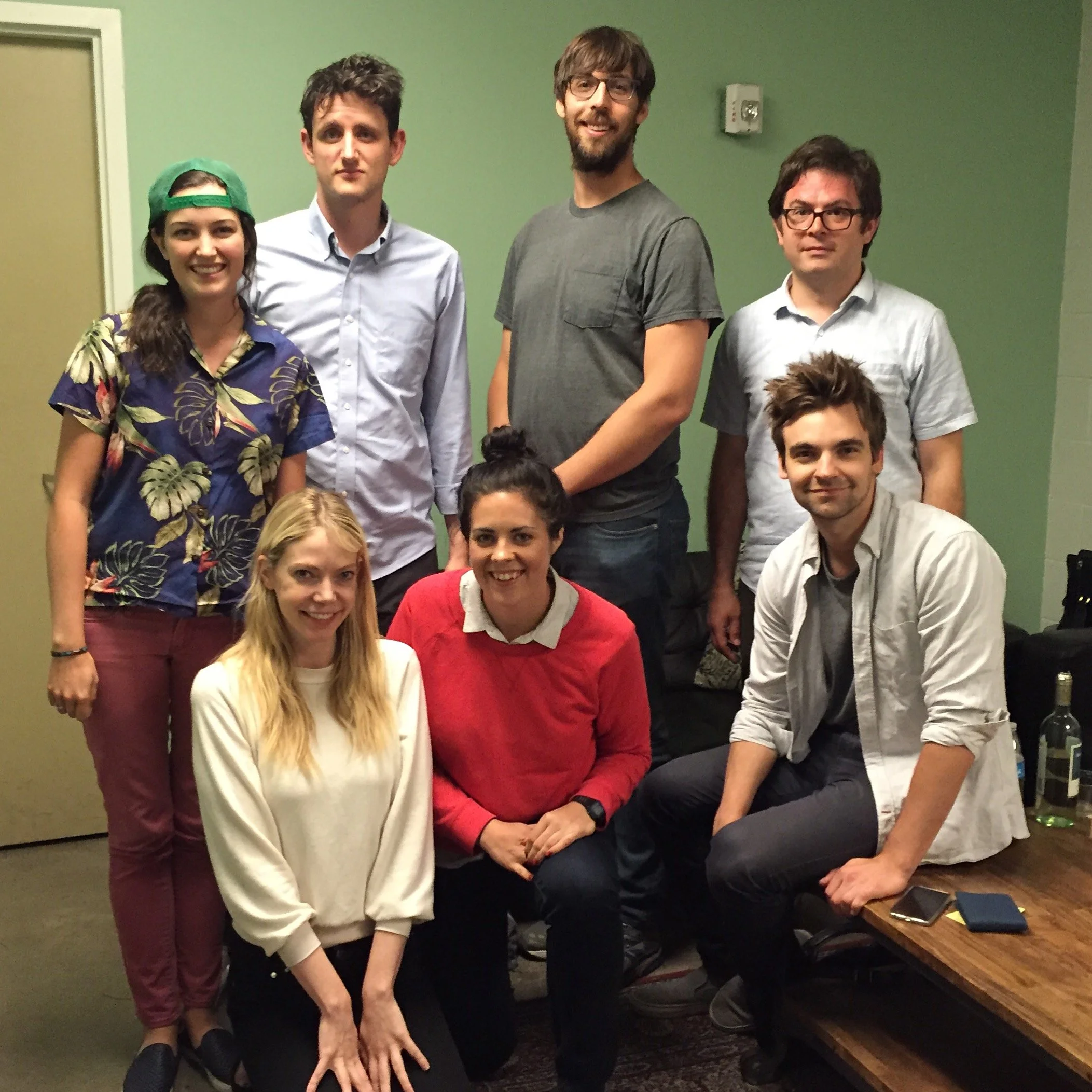 Group of five smiling young adults standing and sitting together in an indoor setting, including a woman wearing a green backwards cap and colorful shirt, along with four men, one with glasses and a beard, another with a gray T-shirt, one in a white shirt, and one in a red sweater with white collar.