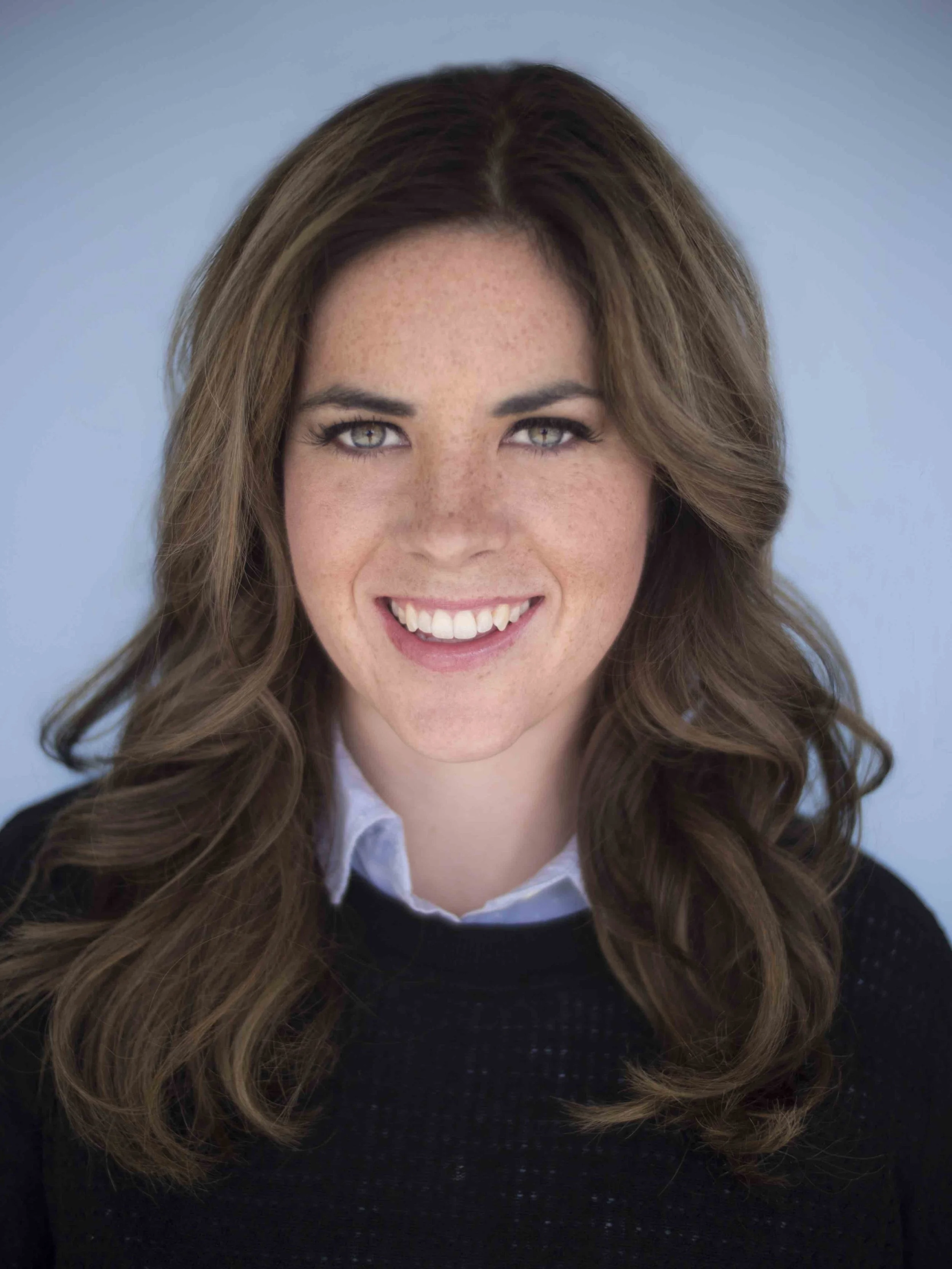 Close-up of a woman with long, wavy brown hair and blue eyes, smiling, against a plain background.