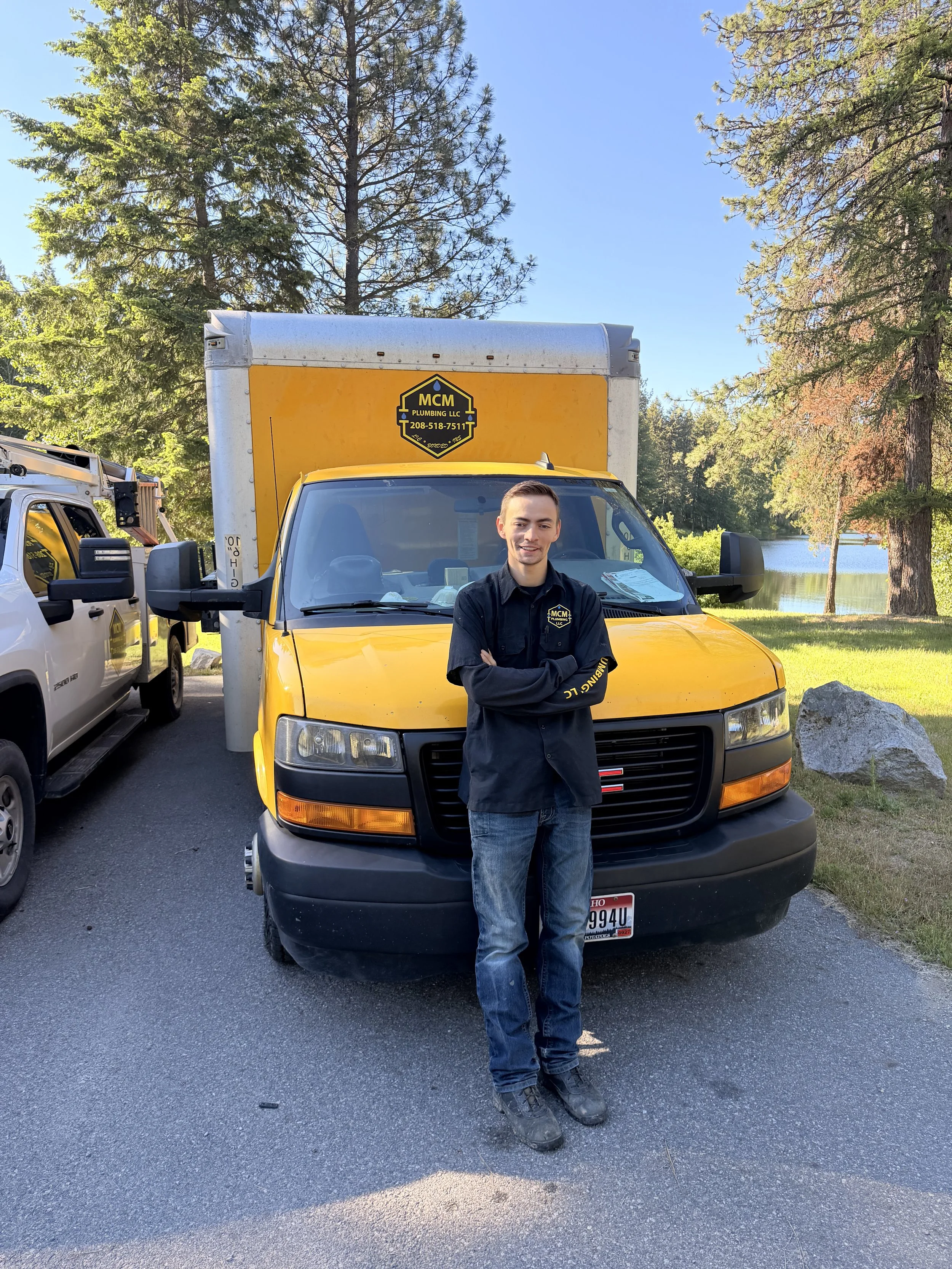 A young man in a black uniform with a logo on the chest and sleeve, standing with arms crossed in front of a yellow service truck with an MCM Plumbing LLC sign. The scene is outdoors near a lake, with trees and blue sky.