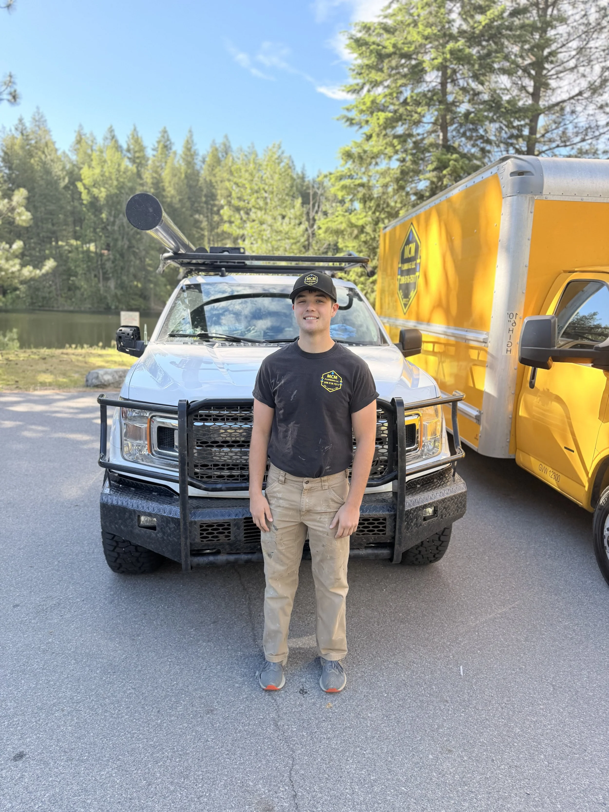 Young man standing in front of a pickup truck and yellow service truck near a lake with trees and blue sky in the background.