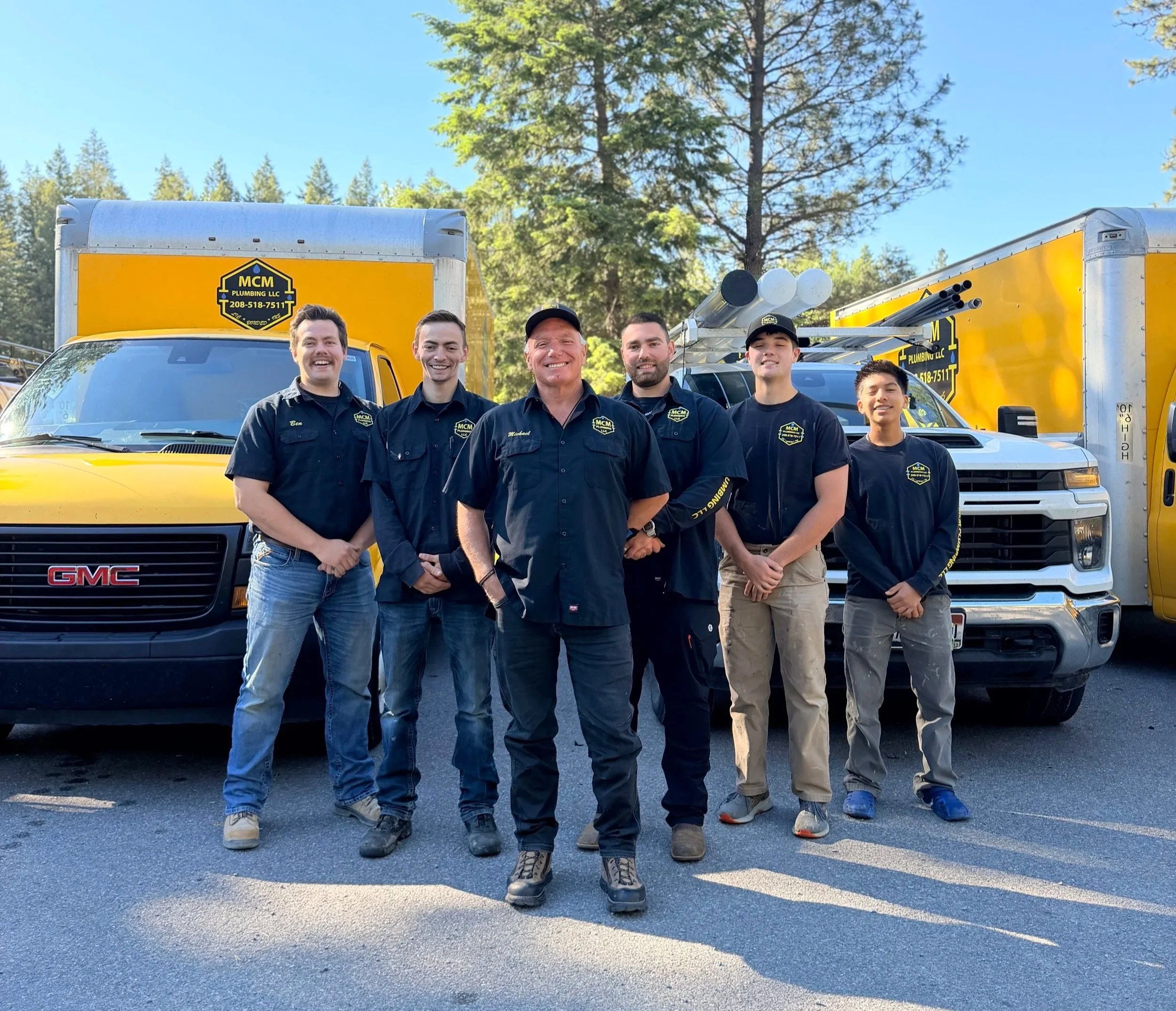 Group of five male workers standing in front of yellow service trucks with ladders, outdoors in a sunny, wooded area.