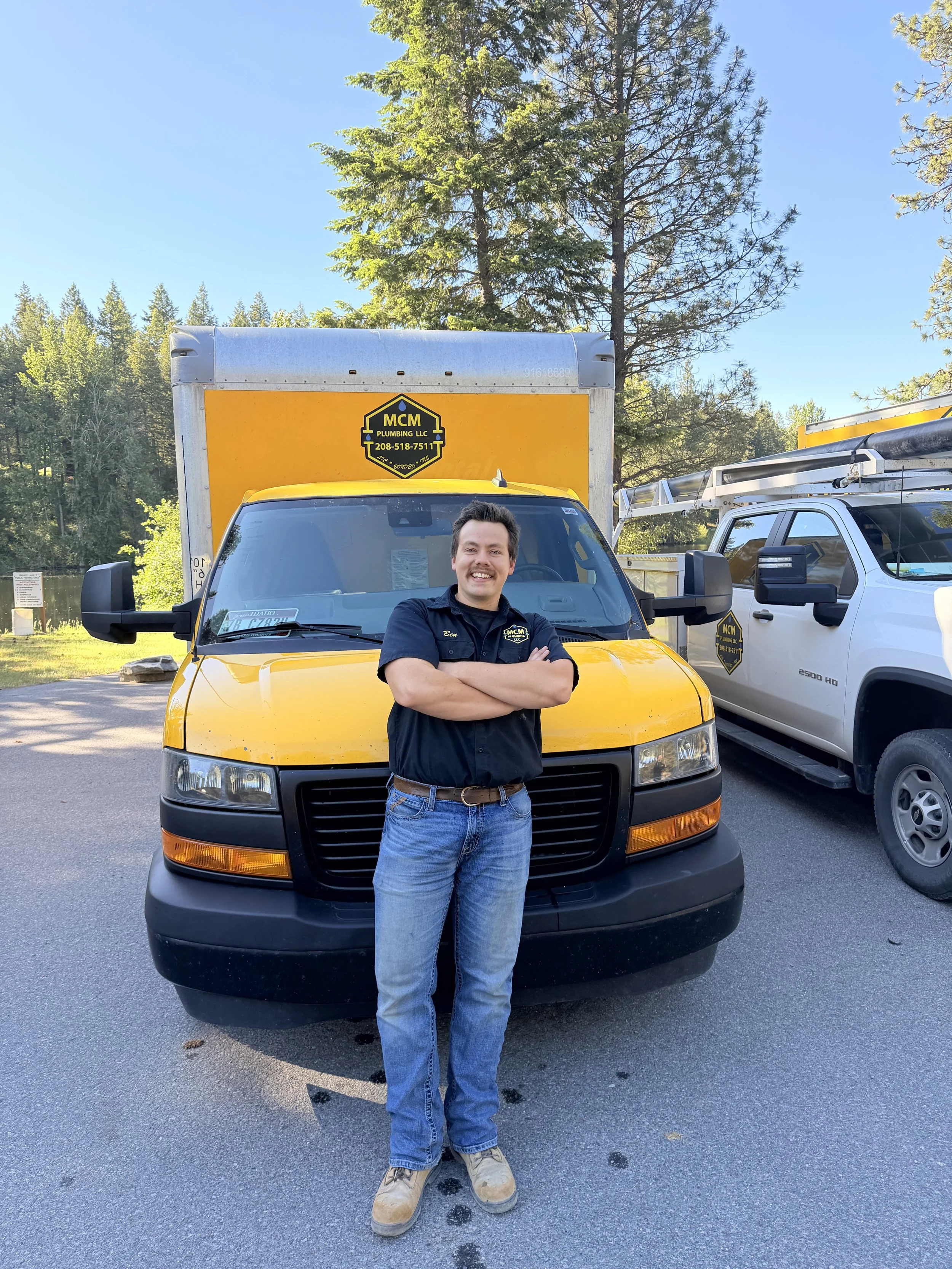 A man stands with his arms crossed in front of a yellow utility truck and a white service pickup truck in an outdoor parking lot surrounded by trees under a blue sky.