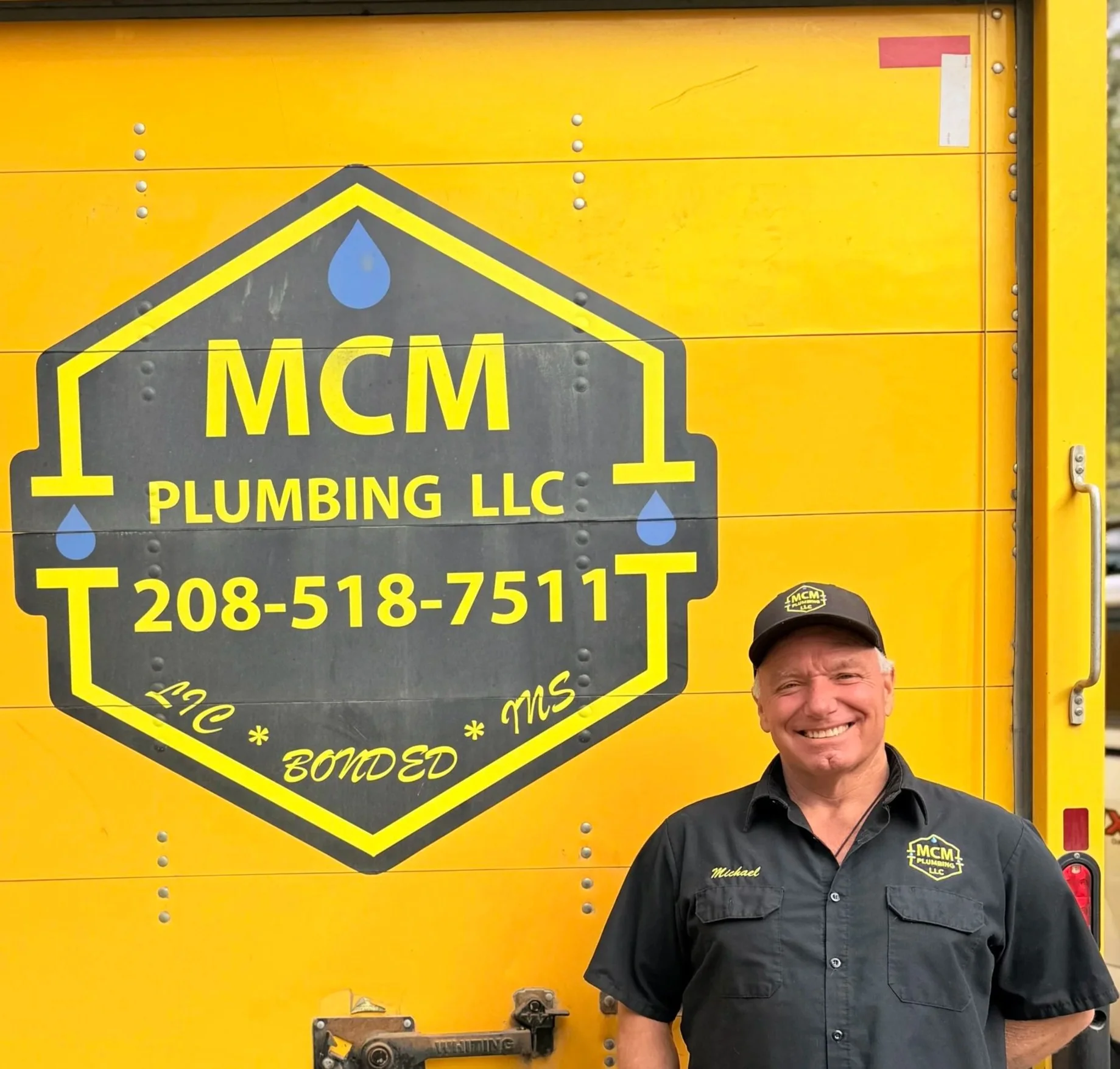 A man smiling and wearing a black uniform with the name Michael and a logo for MCM Plumbing LLC, standing in front of a yellow MCM Plumbing LLC truck with the company's logo, contact information, and blue water droplets design.