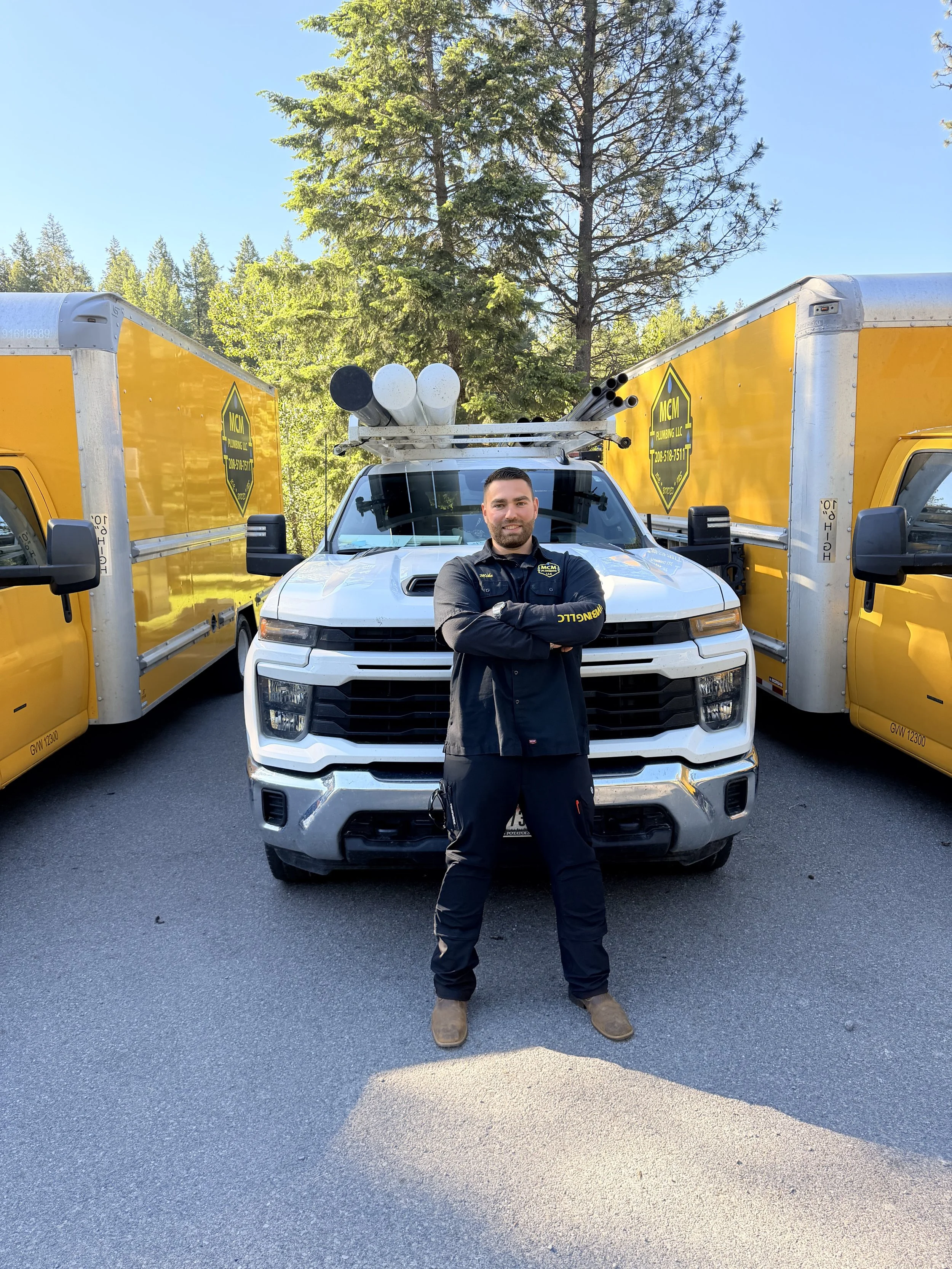 A man stands confidently in front of a white pickup truck with two yellow utility trucks on either side. The man is wearing black work clothes with yellow lettering and brown boots, and has his arms crossed. The background features tall evergreen trees and a clear blue sky.