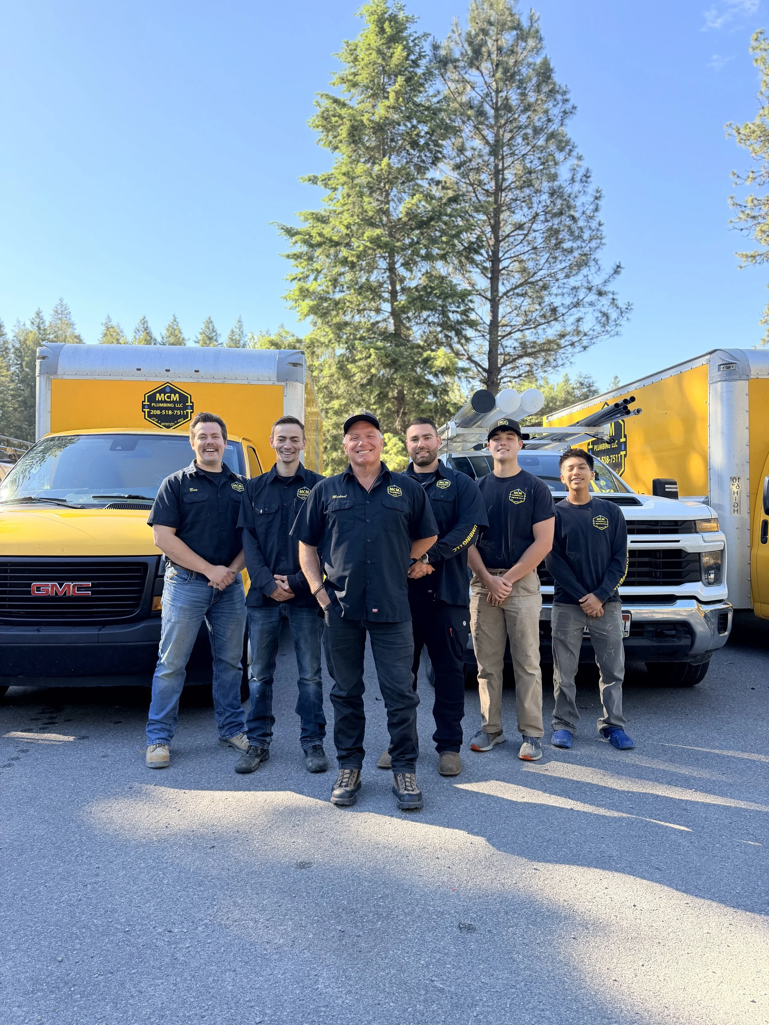 Group of six plumbing technicians standing in front of two yellow MCM Plumbing LLC service trucks outdoors, with trees and a clear blue sky in the background.