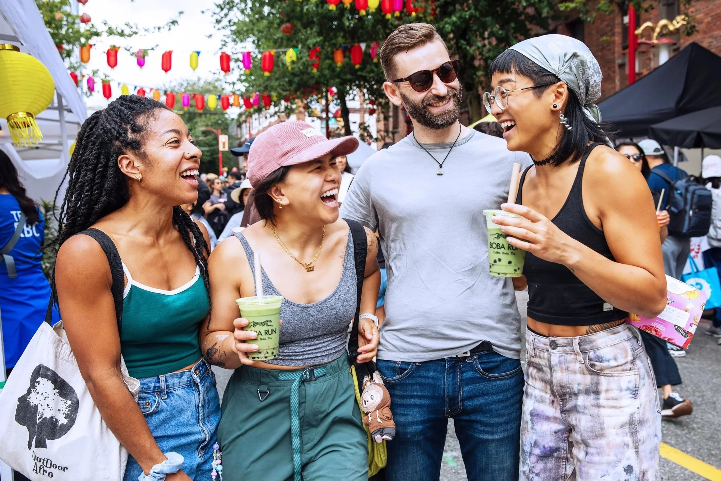 A group of five diverse friends smiling and chatting at an outdoor festival, with colorful lanterns hanging overhead and people in the background.