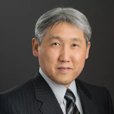 Portrait of a middle-aged Asian man with gray hair wearing a black pinstripe suit and a striped tie, against a black background.