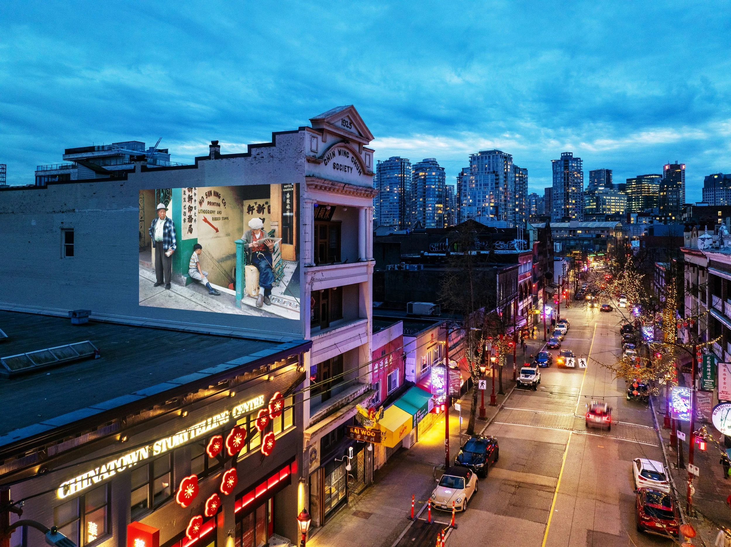 A city street scene at dusk with skyscrapers in the background and a Chinatown sign on a building in the foreground. The street is lined with shops, neon signs, and some parked cars, with trees decorated with lights.