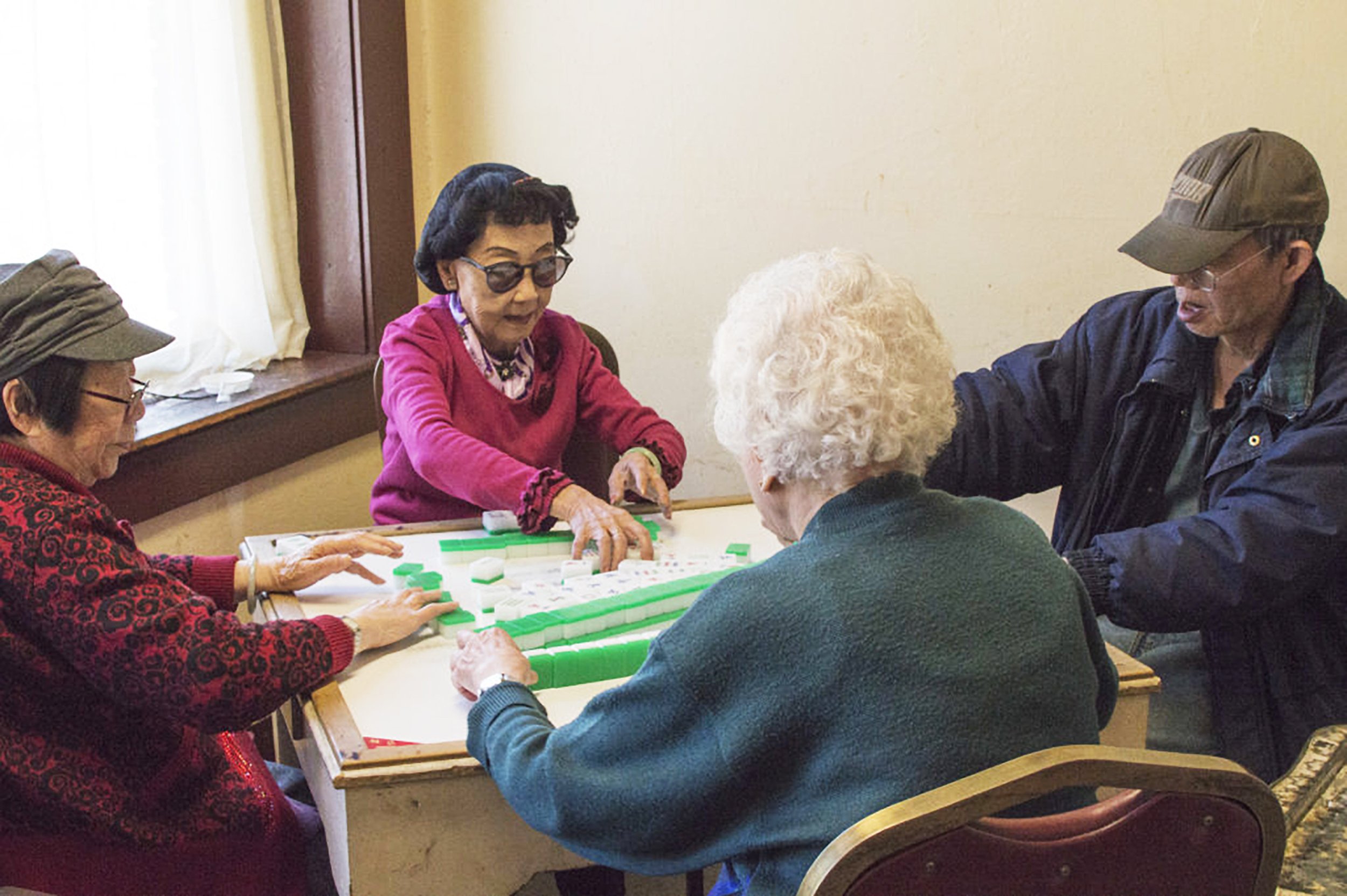 Group of elderly people playing a tile-based game at a table in a community room.