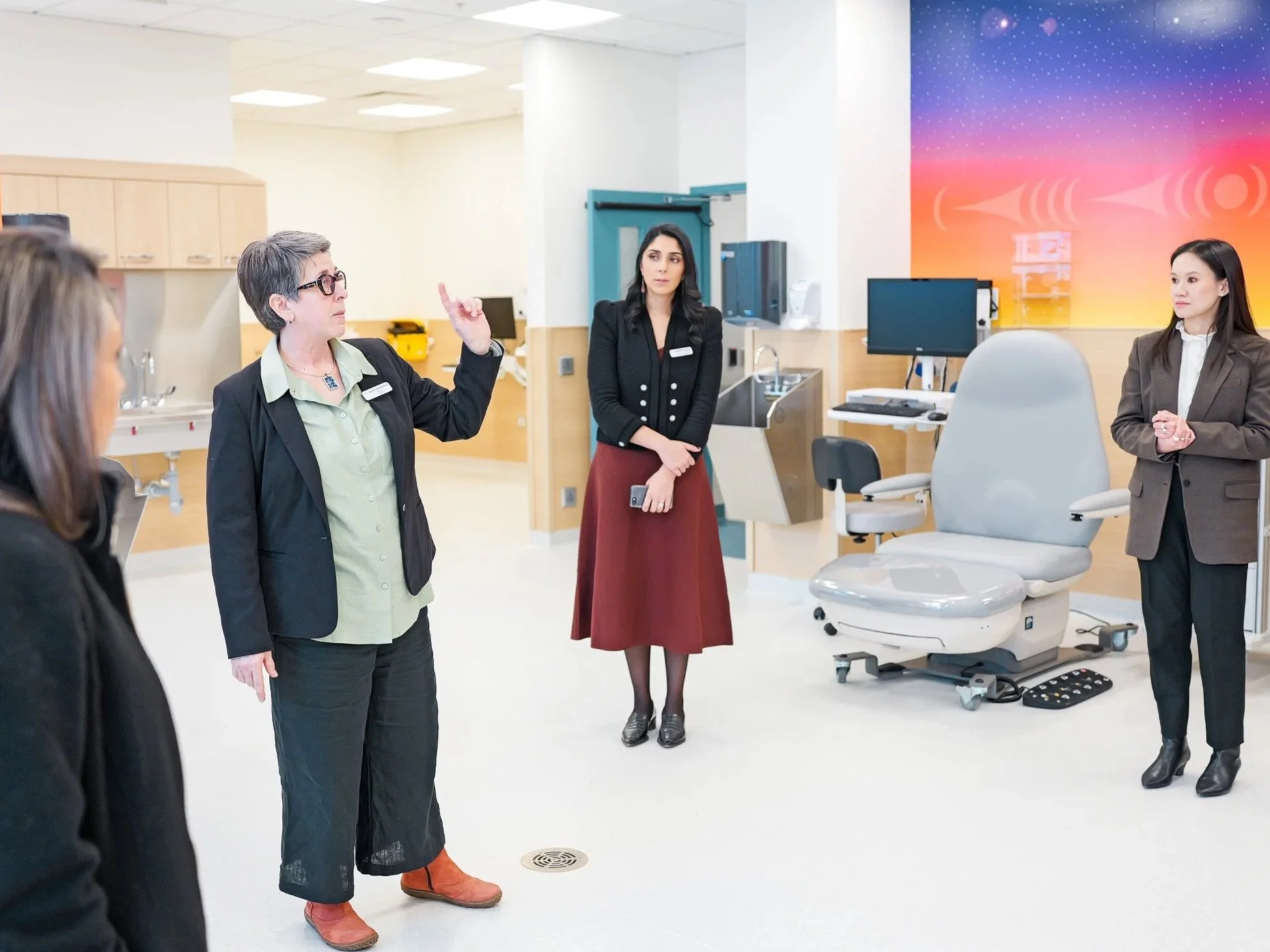 Group of women in professional attire having a discussion in a medical or hospital setting with medical equipment in the background.