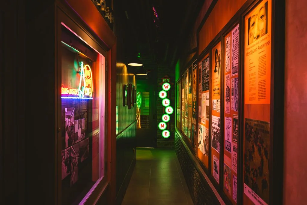 A dimly lit, colorful hallway with neon signs and framed posters on the walls, including a vertical illuminated sign that spells out "WELCOME" in green and white lights.