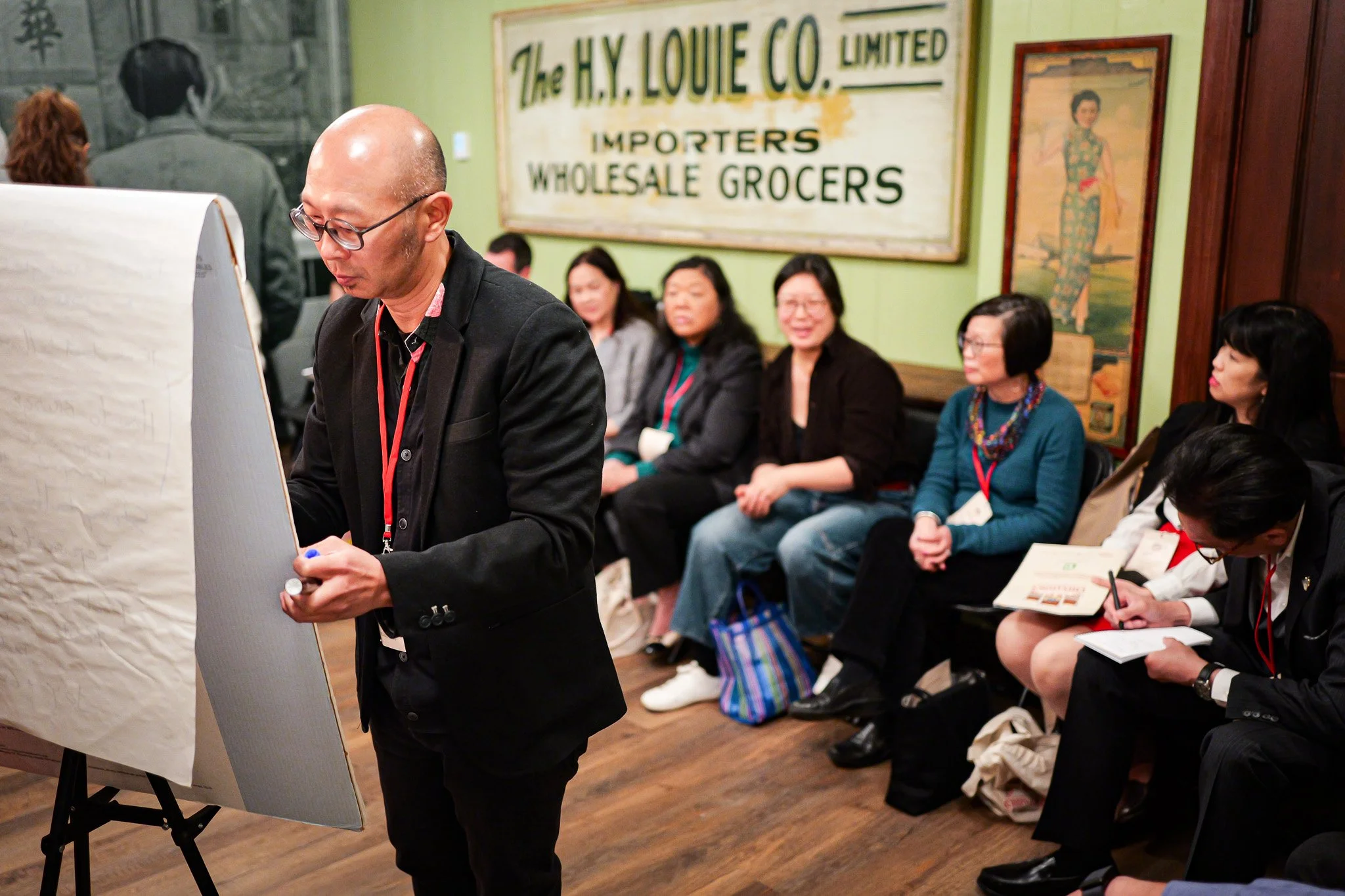 A man with glasses and a bald head dressed in black is writing on a large paper sheet on a stand. Behind him, there is a group of women seated, some of them taking notes or holding papers. The background features a large vintage sign reading 'The H.Y.Louie Co. Ltd., Importers Wholesale Grocers' and some framed artwork.