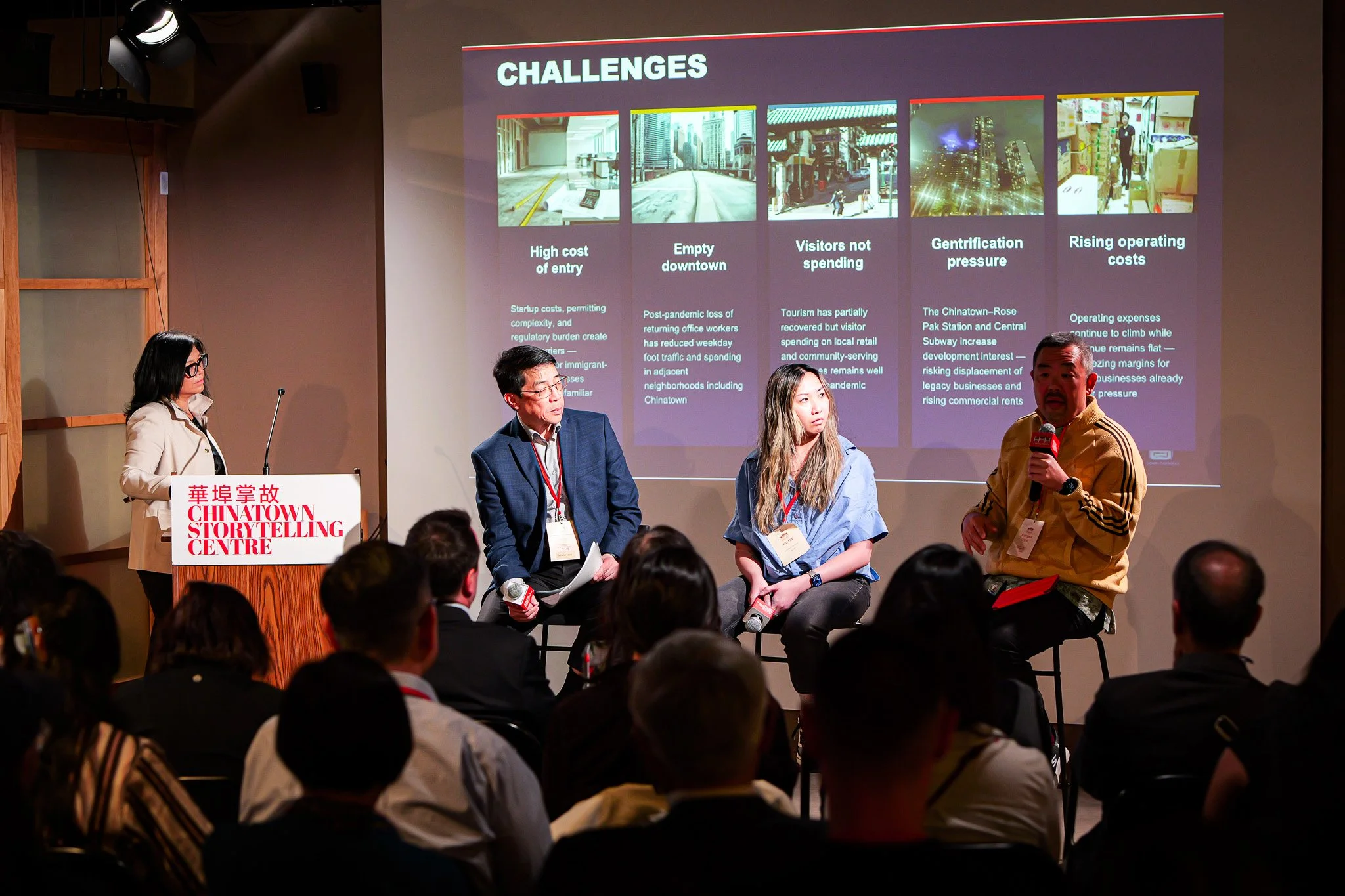People seated on stage during a panel discussion at the Chinatown Storytelling Centre. A large presentation slide behind them lists challenges such as high entry costs, empty downtown, visitors not spending, gentrification pressure, and rising operating costs.