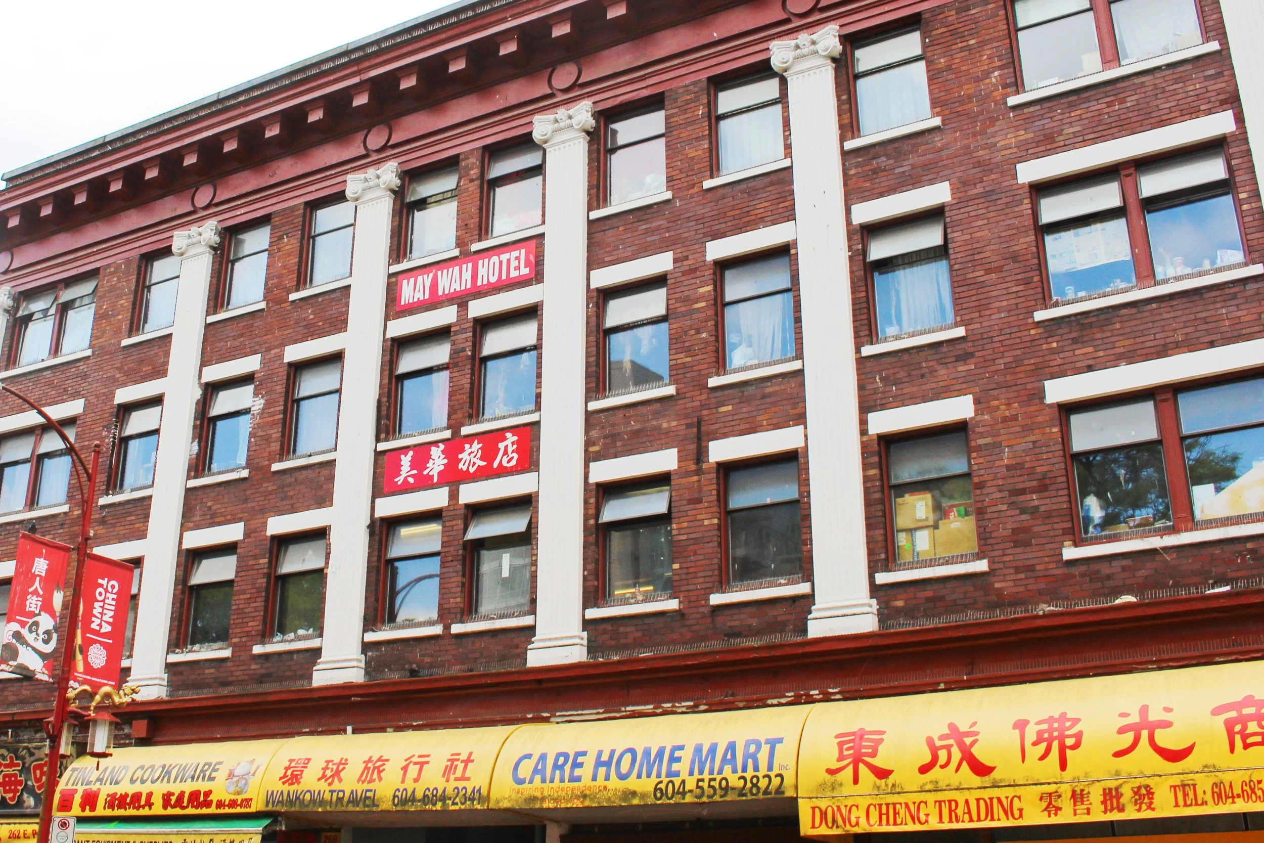 The image shows the exterior of a multi-story brick building with a sign that reads 'MAY WAH HOTEL' and Chinese characters below. The building has several windows with some displaying items inside. There are also red banners with Chinese characters hanging on poles in front of the building, and a yellow awning with additional Chinese and English text.