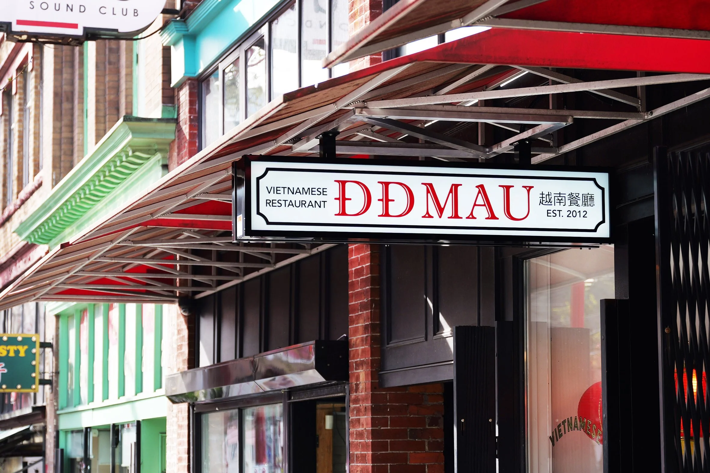 Signboard for Vietnamese restaurant named 'DE MAU' with establishment year 2012, hanging outside a brick building with green and red awnings.
