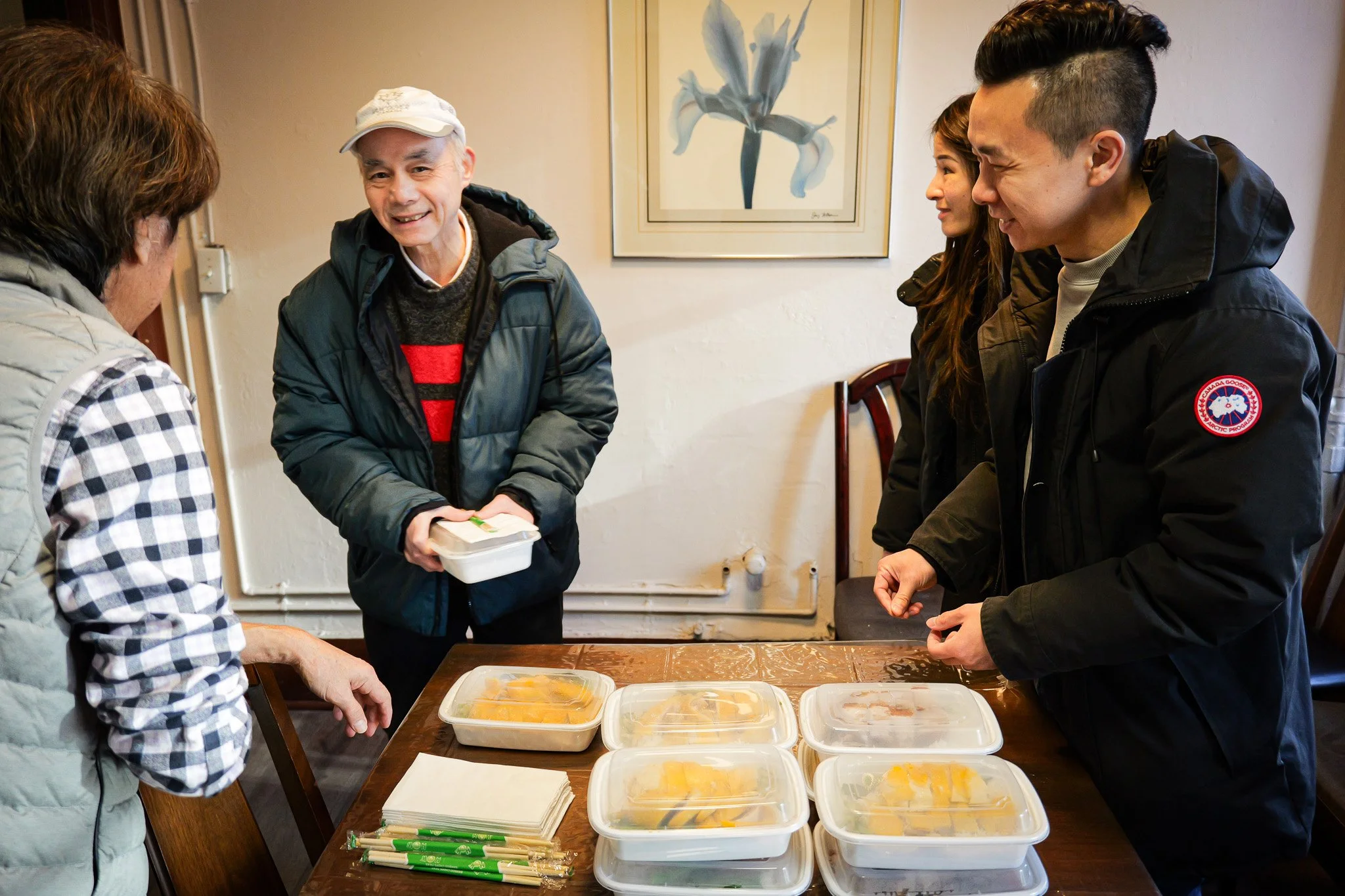 A group of five people standing around a wooden table with food containers and chopsticks, engaging in a conversation. One man in the center, wearing a dark jacket and a white cap, is smiling and holding a plastic container. The others are smiling and listening. A framed flower painting hangs on the wall behind them.
