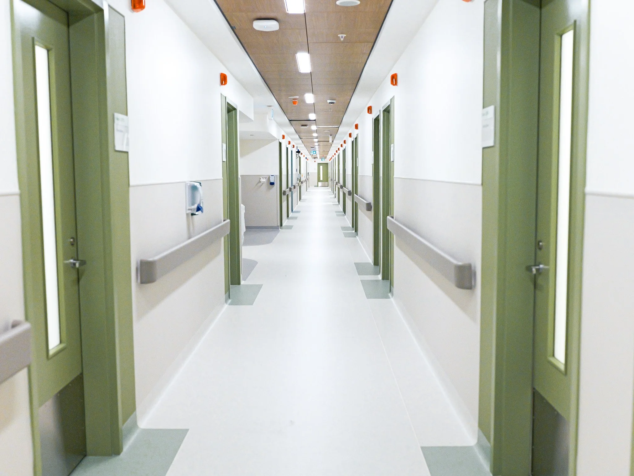 A hospital corridor with multiple elevator doors on each side, light-colored walls, and a ceiling with fluorescent lighting.
