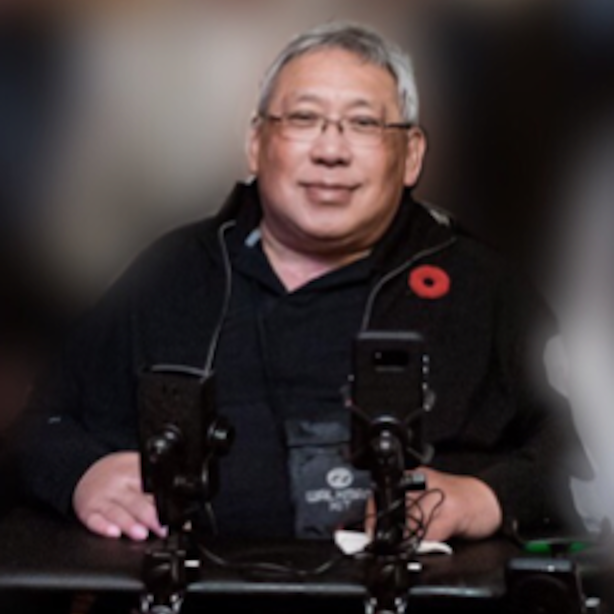A smiling man with glasses and gray hair sitting at a table with two mounted phones, wearing a black jacket with a red poppy pin.