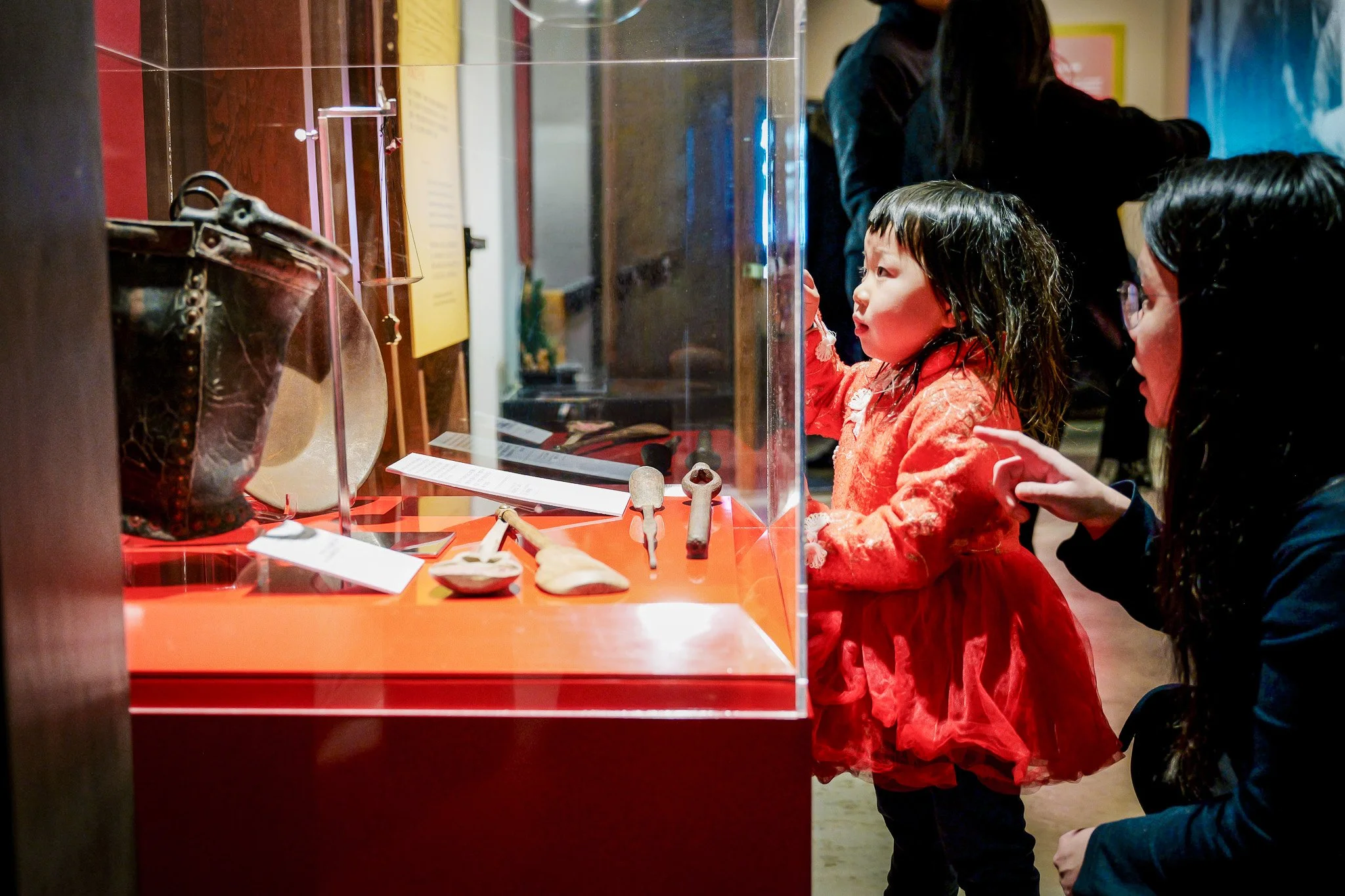 A young girl in an orange dress and an adult woman with sunglasses look at ancient artifacts and fossils displayed in a glass case at a museum.