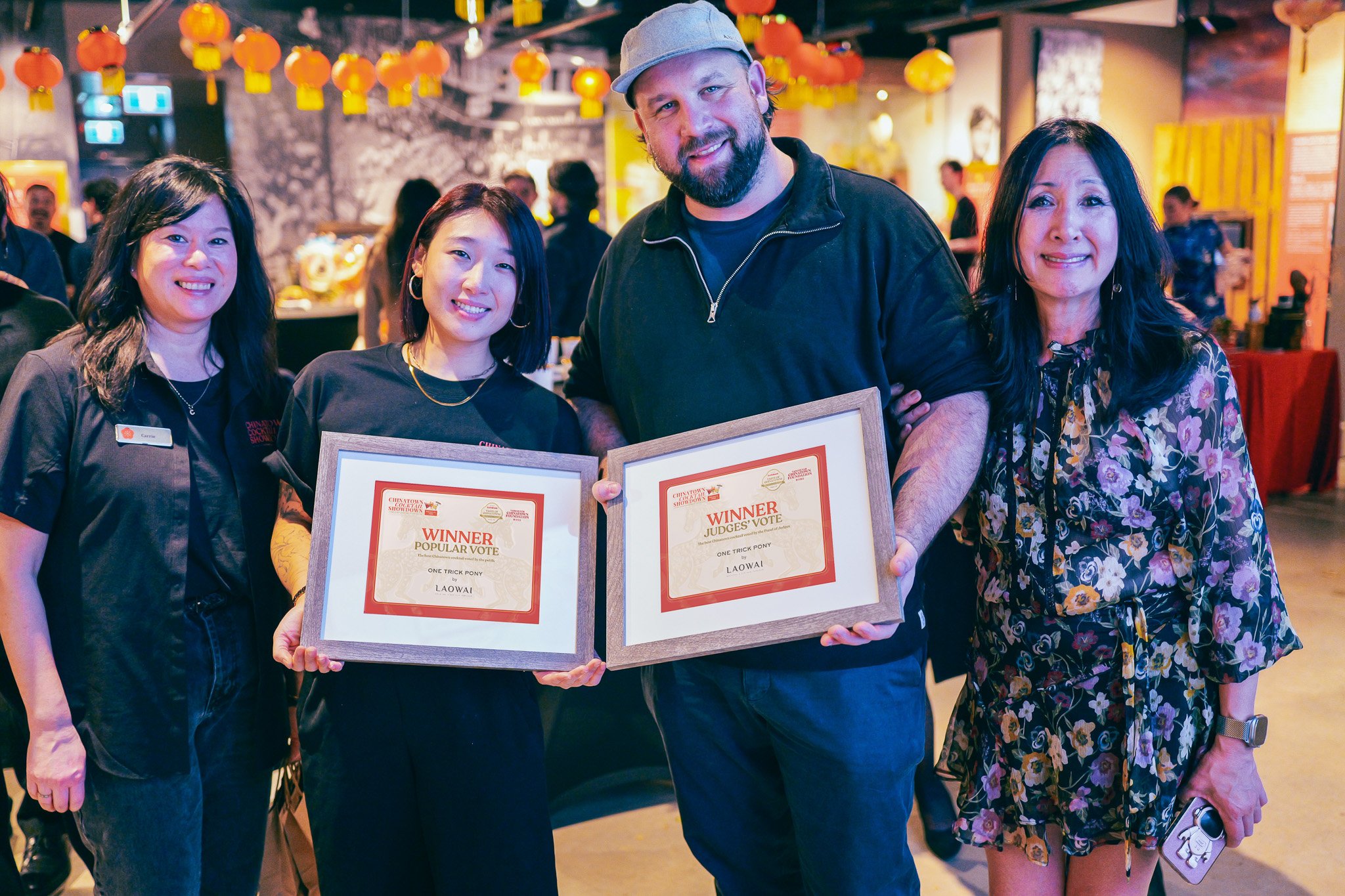 Group of four people smiling and holding awards at an indoor event with Chinese lanterns hanging from the ceiling.