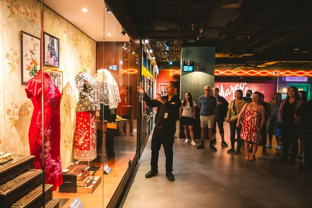 A museum tour guide is explaining traditional Chinese dresses displayed in a glass case to a group of visitors inside a museum.