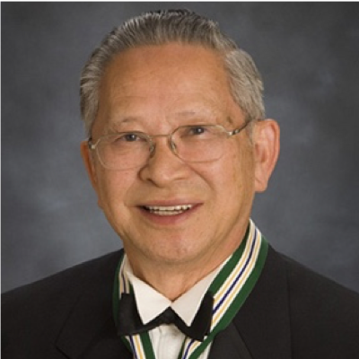 A portrait of an elderly man with gray hair and glasses, smiling, dressed in a suit with a black bow tie and wearing a medal around his neck against a gray background.