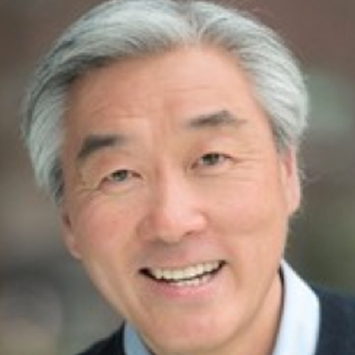 Close-up of a smiling middle-aged man with gray hair, wearing a dark jacket and light shirt.