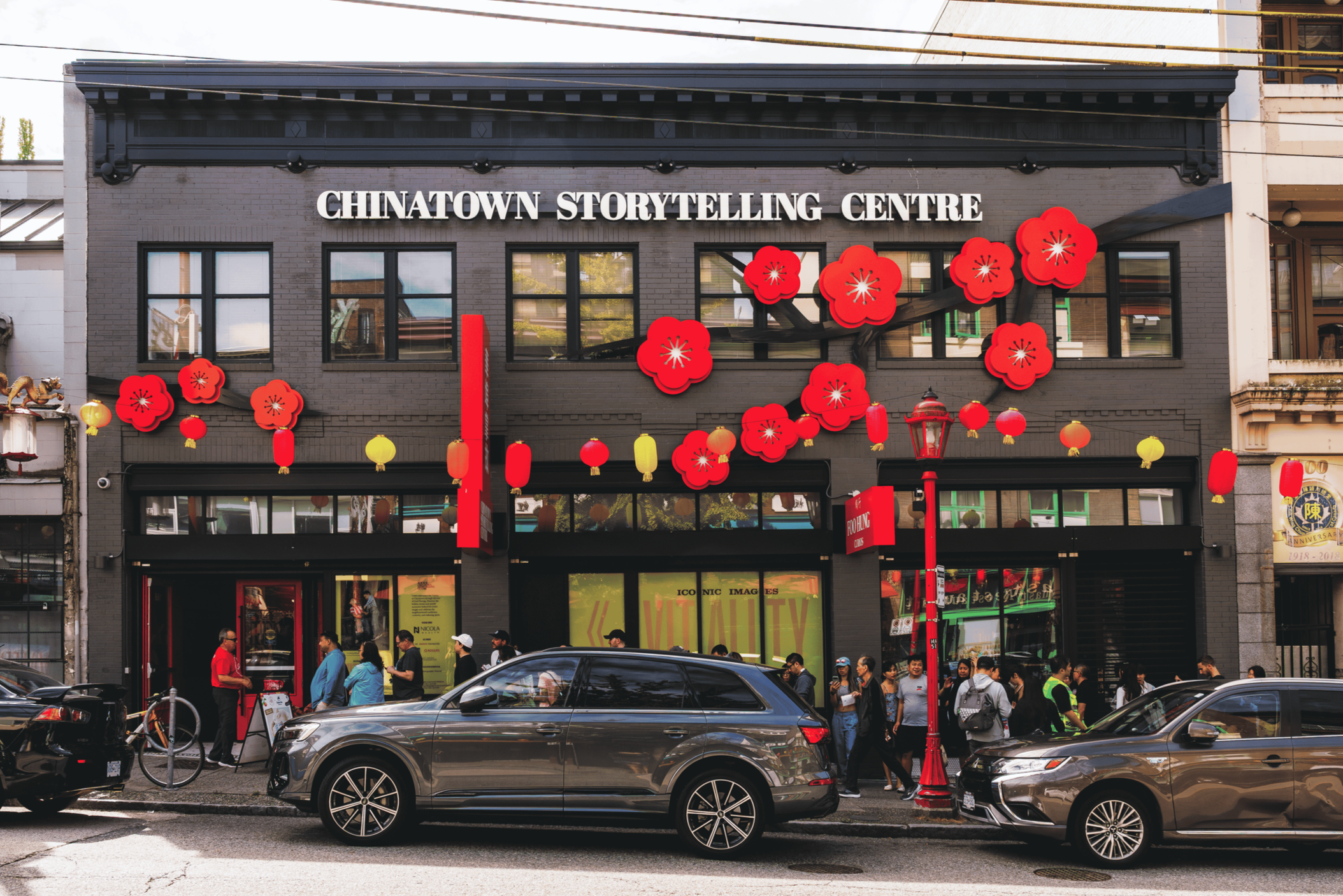 The facade of a building labeled 'Chinatown Storytelling Centre' decorated with red and yellow lanterns and cherry blossom decorations, with people walking and cars parked in front.