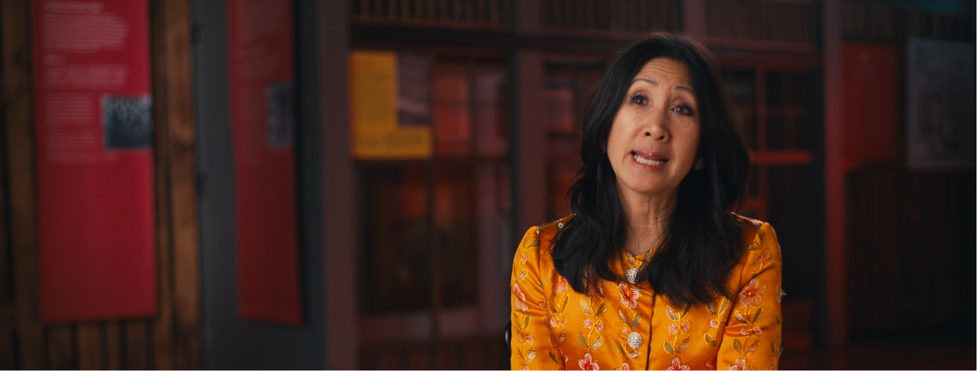 A middle-aged woman with long black hair and a floral orange dress, speaking in front of bookshelves in a room with red wall art.