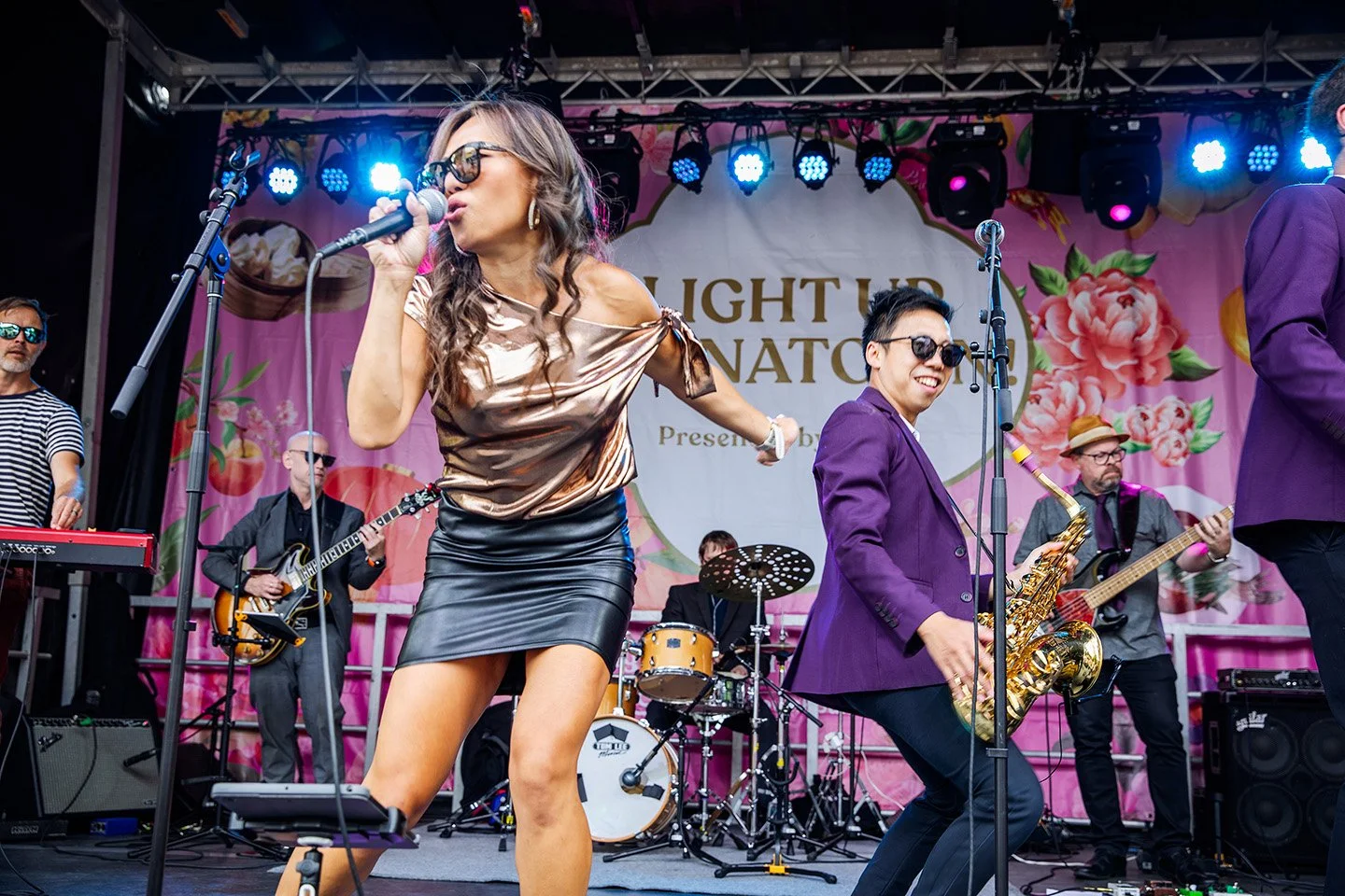 A band performing on stage at an event, with a woman singing and a man playing saxophone, surrounded by other musicians, against a pink floral backdrop.