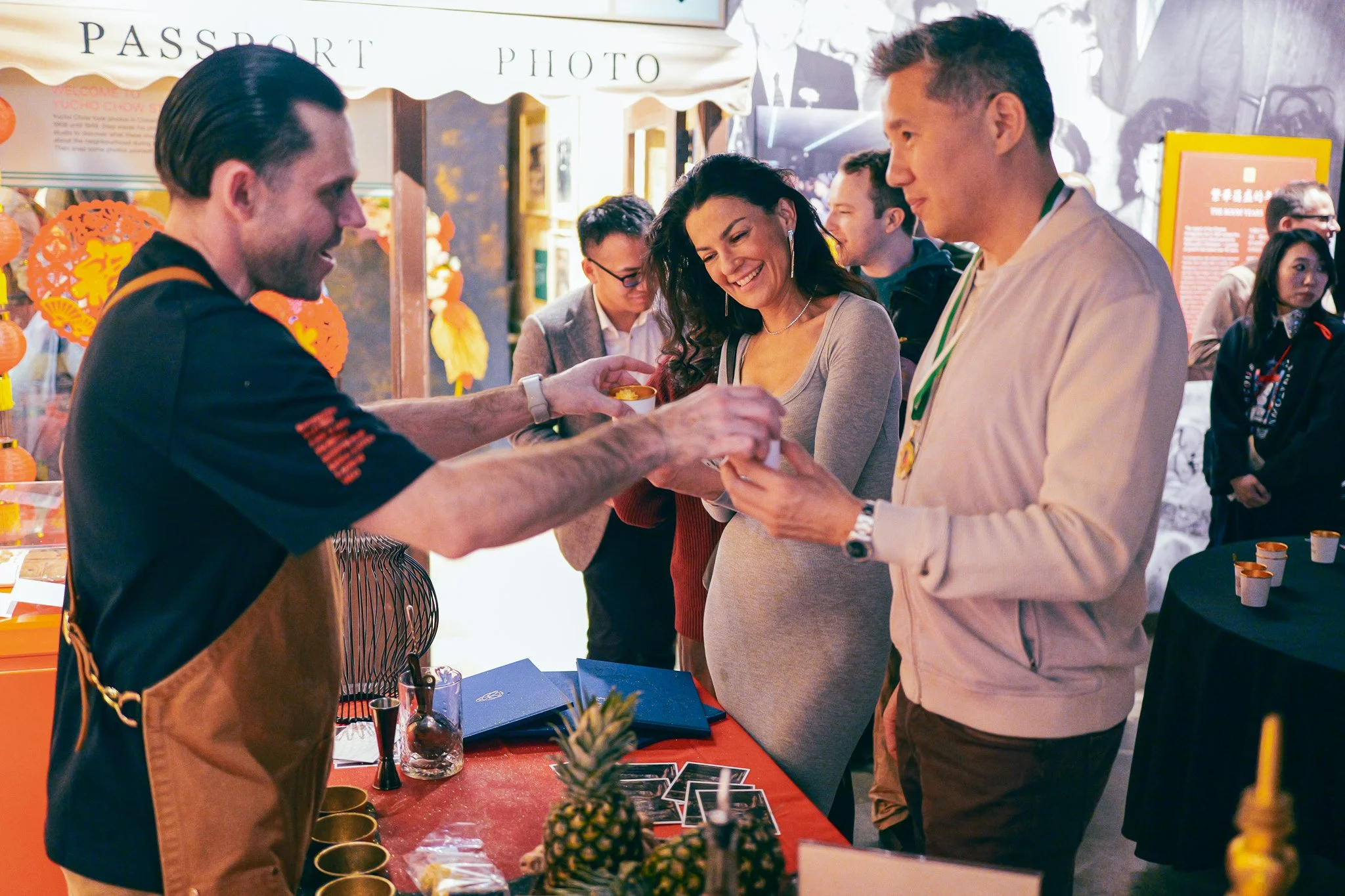 A group of people at a booth with a red tablecloth, exchanging a small item. The booth has a pineapple and various small cups on the table. In the background, there are signs and other people at the event.