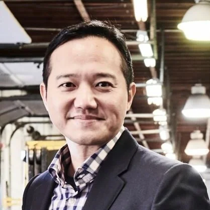 A man in a business suit smiling in an industrial or warehouse setting.
