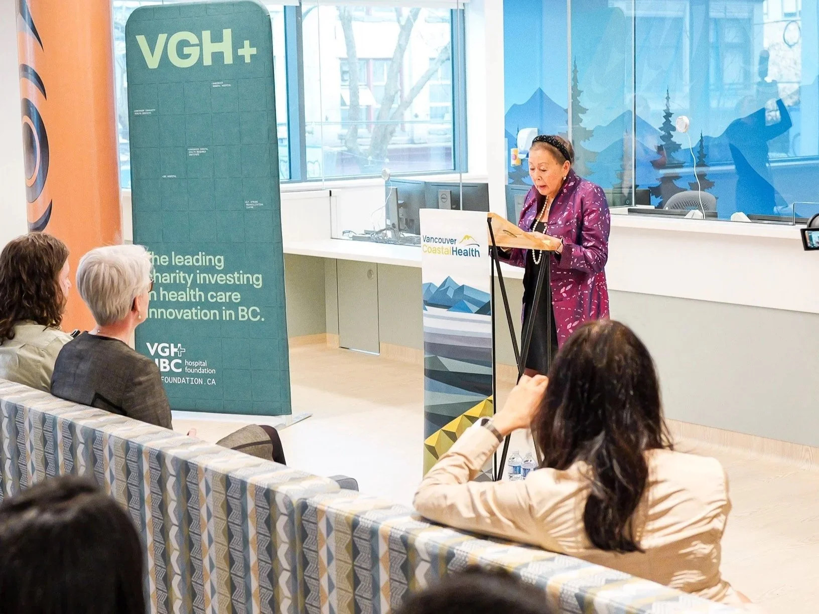 A woman in a purple dress with a headband speaking at a podium with a Vancouver Coastal Health banner, with an audience seated in front of her inside a room with large windows showing trees outside.