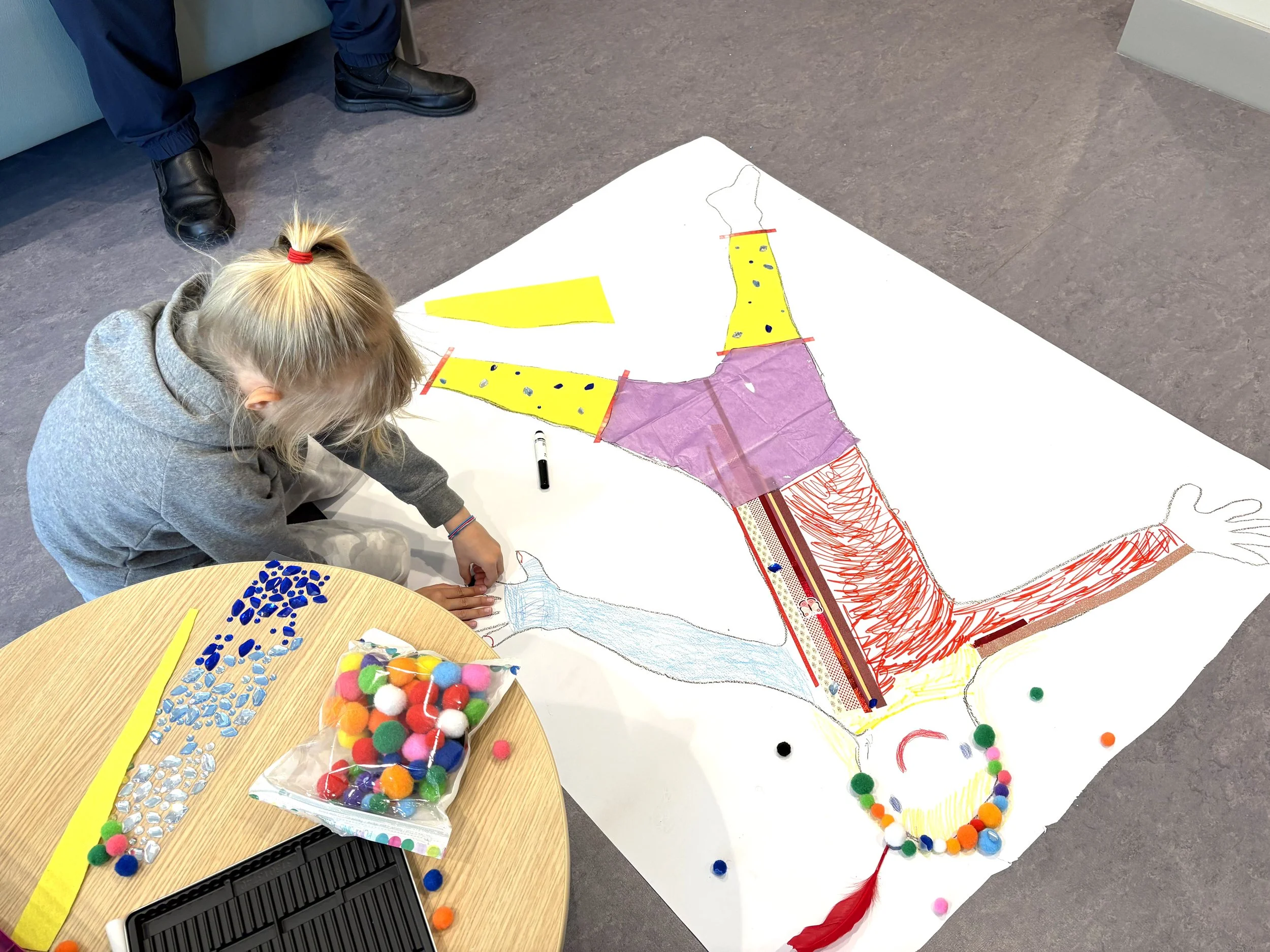 A young girl is working on an art project, creating a large, colorful, paper-based artwork of a dragon on a white poster board. The dragon features yellow, pink, red, and purple sections with various patterns, and is decorated with pom-poms and small blue and transparent gems spread across the surface. There is a container of multicolored pom-poms and a bag of small pom-poms placed on a nearby round table. A person wearing black shoes and navy pants is partially visible standing in the background.