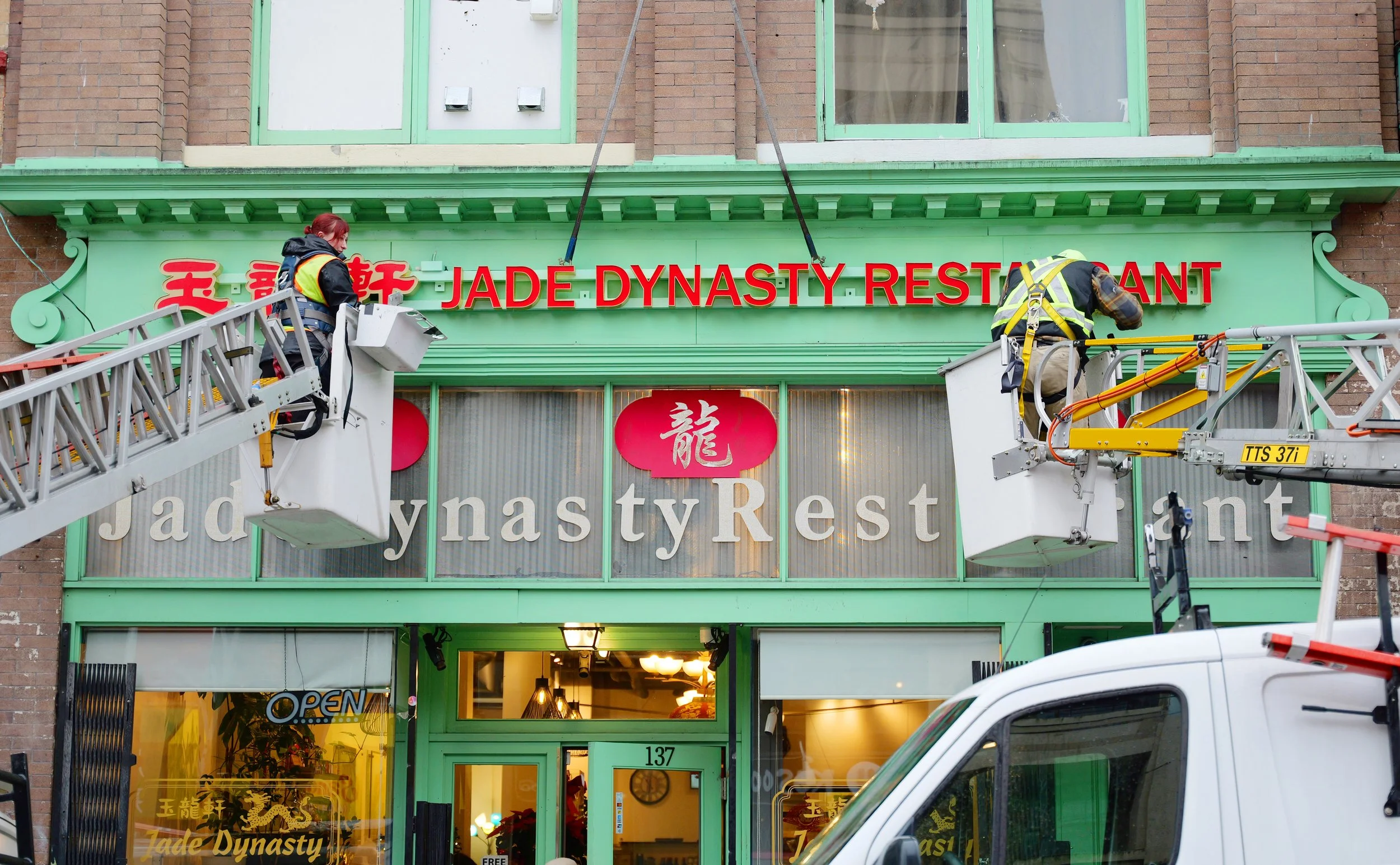 Workers on a lift installation sign for Jade Dynasty Restaurant with Chinese characters, storefront with large windows and open sign.