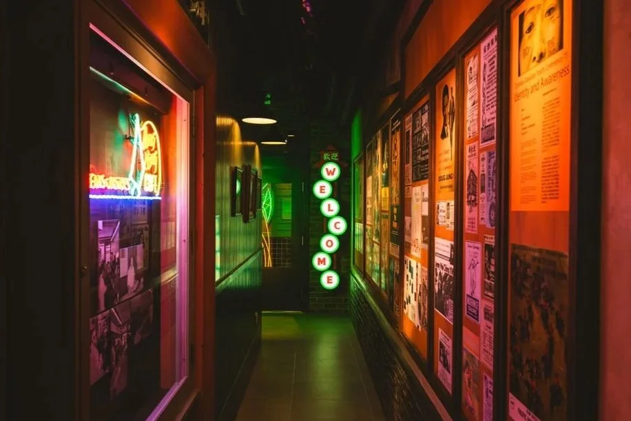 Dimly lit hallway with neon lights spelling 'WELCOME' vertically on a wall, framed pictures and colorful posters on the right wall, and a neon sign on the left, creating a vibrant, retro atmosphere.