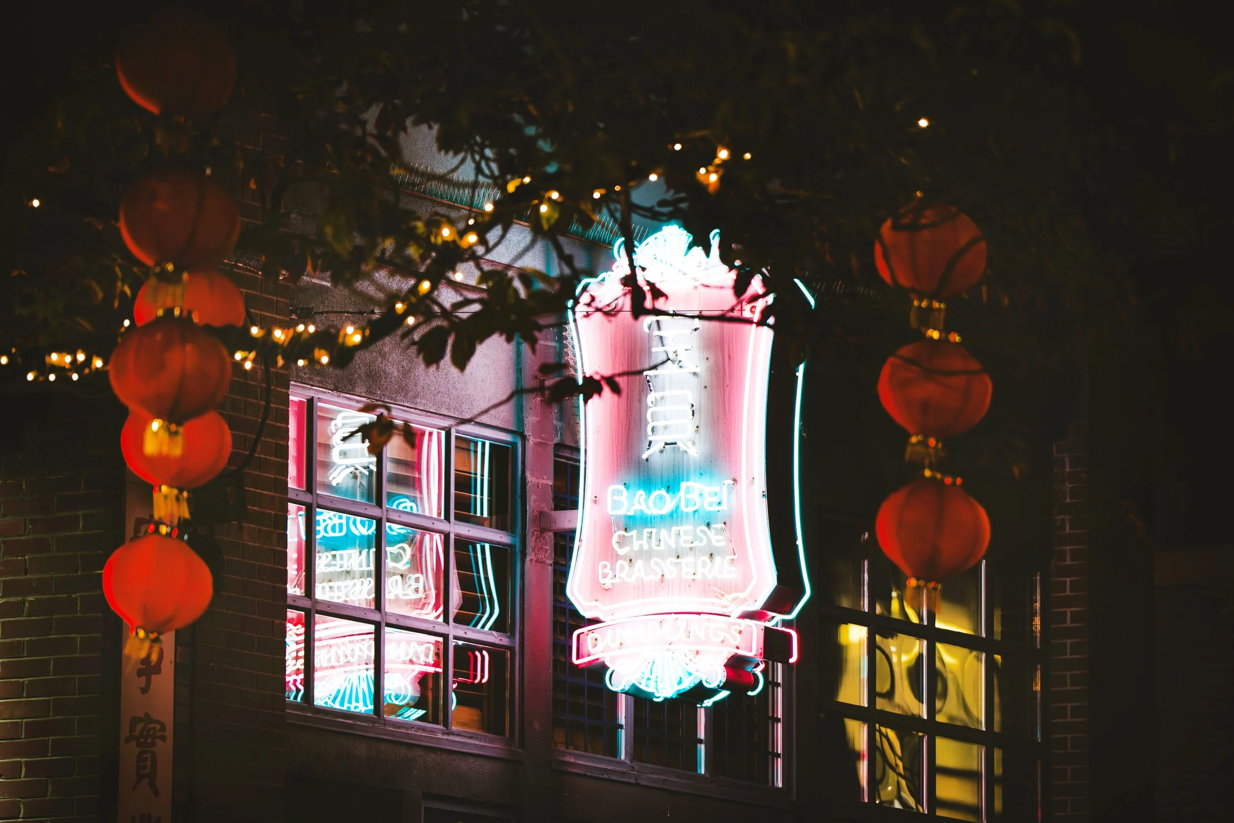 Neon sign for a Chinese brasserie viewed through a window with red paper lanterns and string lights at night.