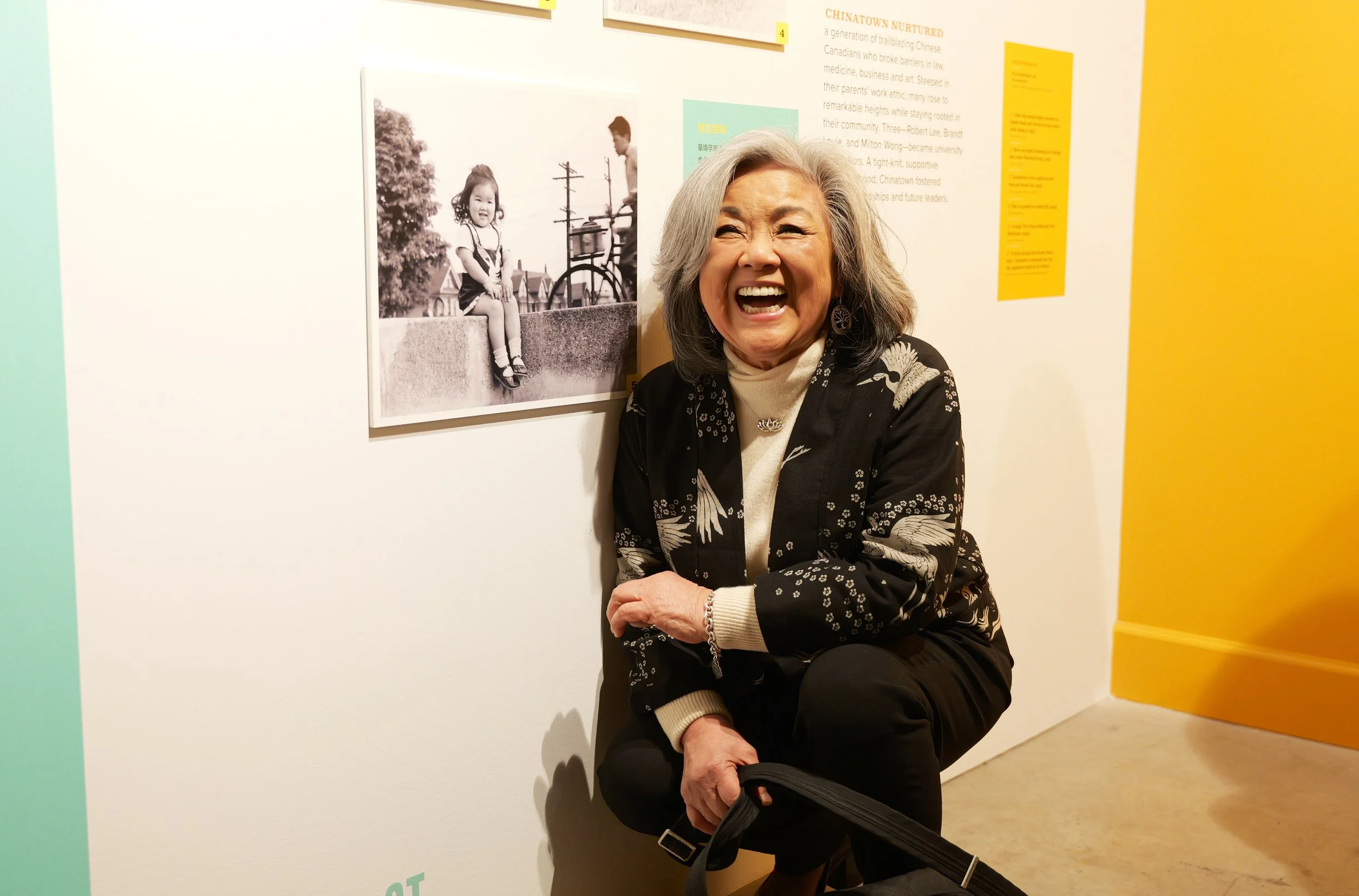 An older woman with gray hair, laughing, sitting on the floor against a white wall in an art gallery. Behind her is a large black and white photograph of a young girl sitting on a ledge and a man with a bicycle. The gallery's wall features colorful panels of text and images.