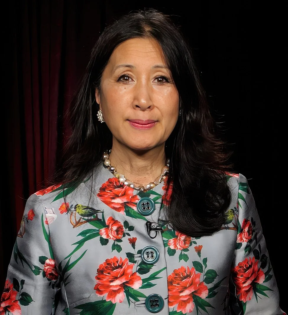 A woman with long black hair, wearing a floral blazer with red flowers, a pearl necklace, and earrings, standing against a dark background.